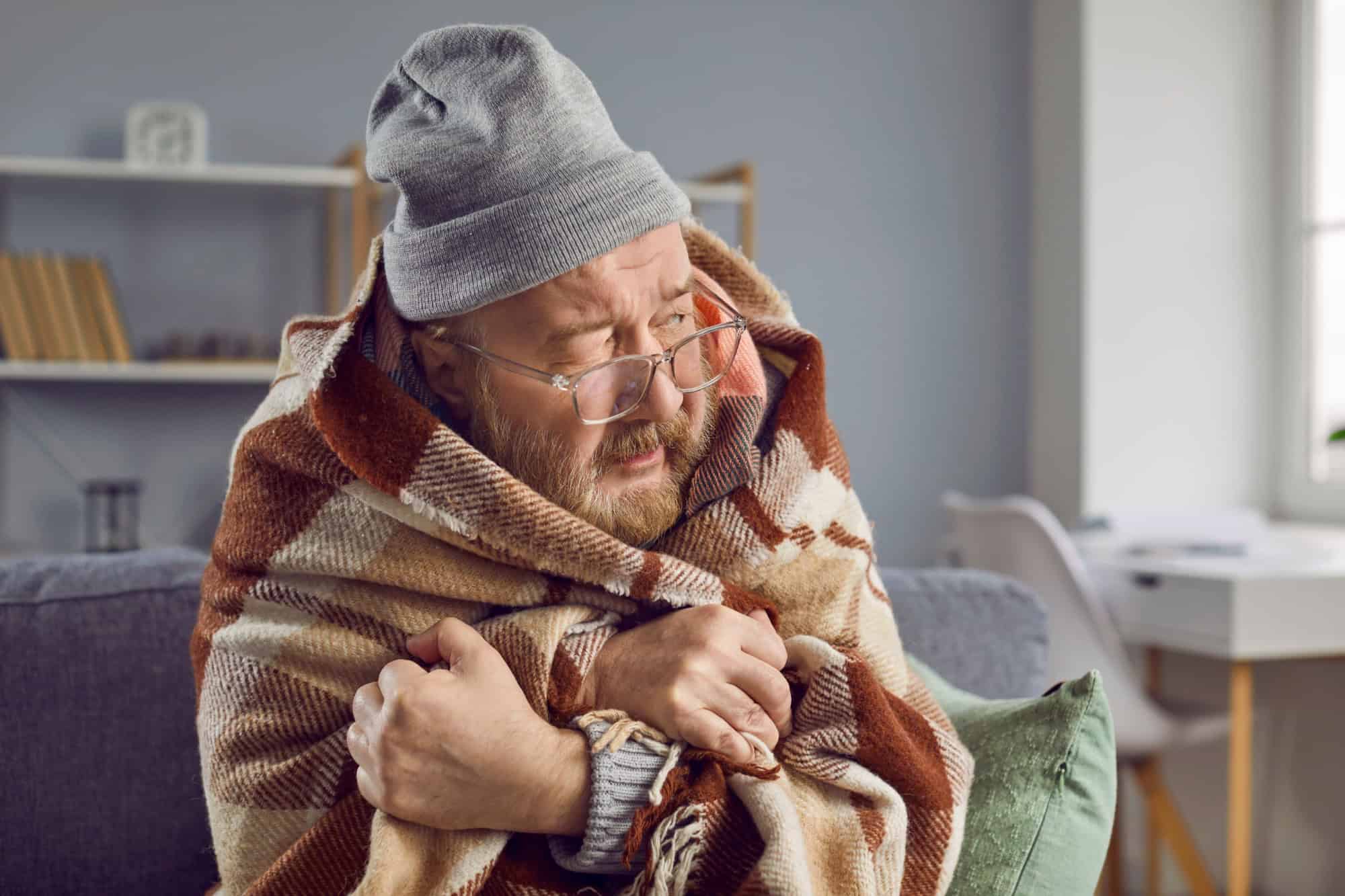 Senior man freezing at home in winter. Elderly man wearing hat and wrapped in blanket sitting on sofa in chilly room with low temperature, in house with bad broken thermostat, in very cold wintertime
