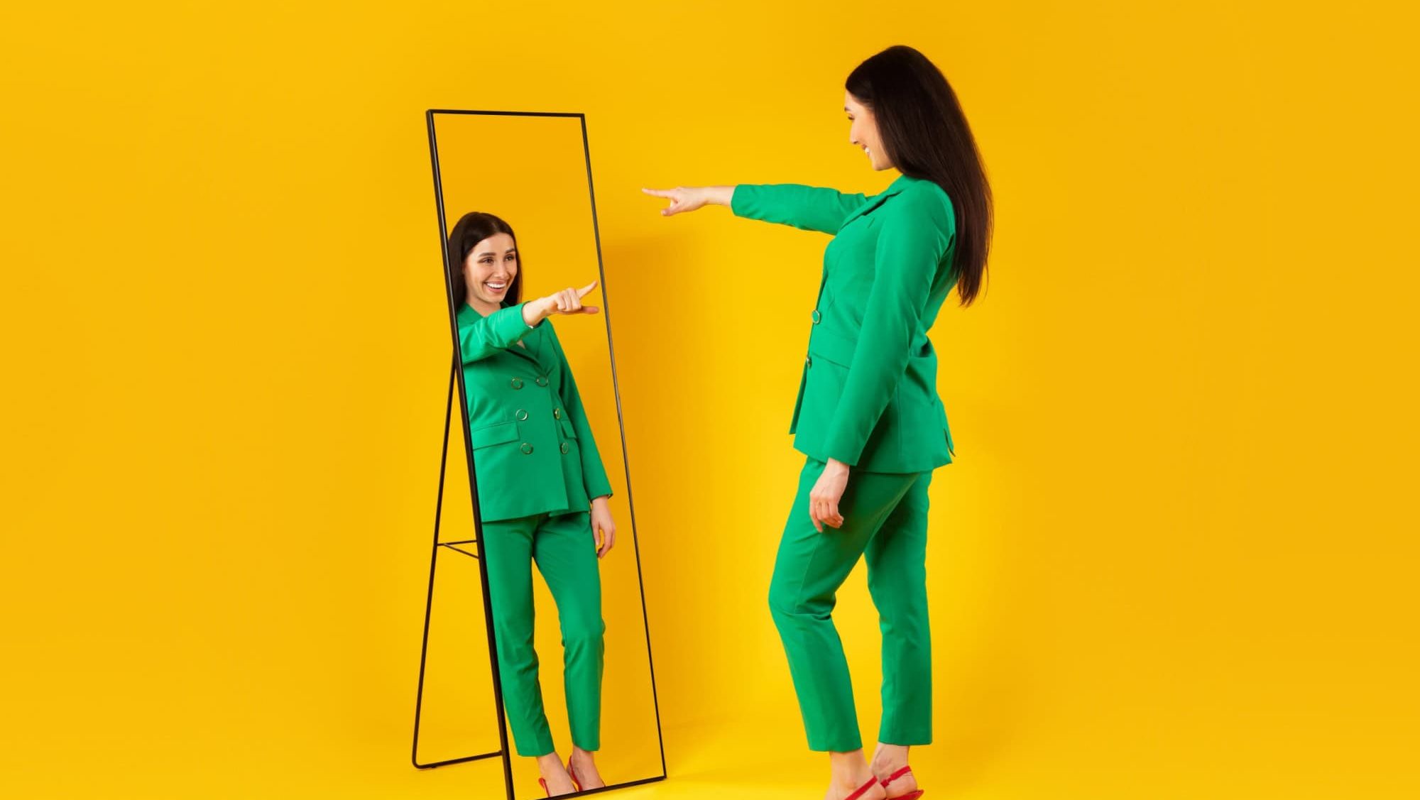 I choose myself. Excited young stylish lady pointing finger at her reflection in mirror, enjoying her new outfit, posing on yellow background, full length, free space. Self confidence concept