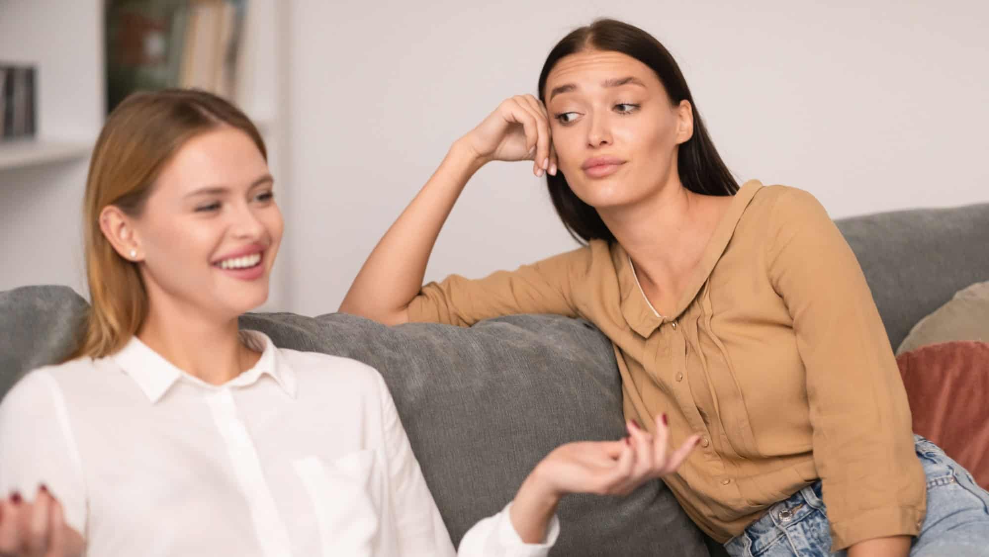 Envy In Friendship. Jealous Lady Listening To Her Cheerful Friend Talking And Bragging About Her Great Life Sitting On Couch At Home, Rolling Eyes During Conversation. Selective Focus