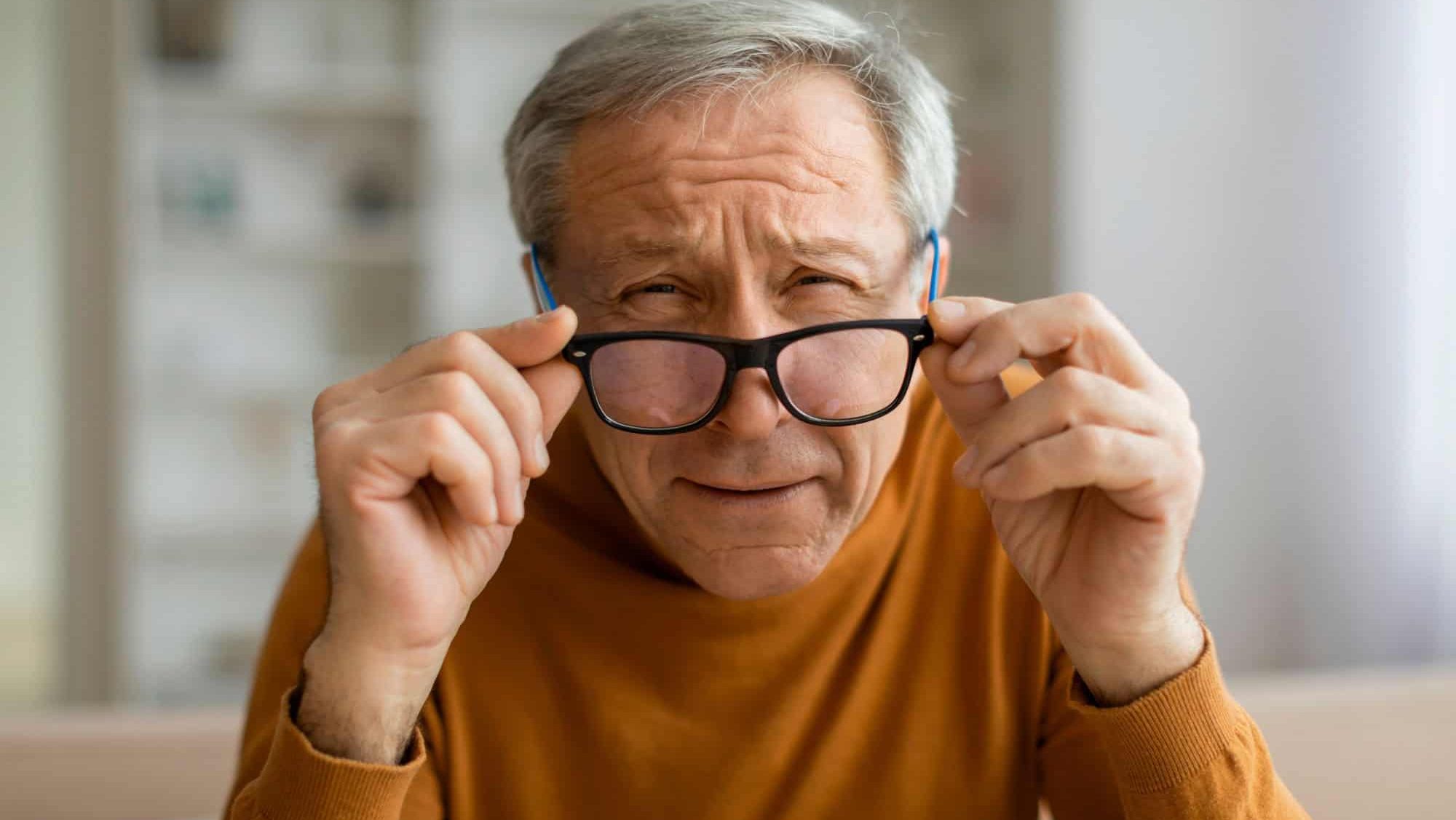 Tired grey-haired elderly man removing eyeglasses and squinting, sitting on couch, home interior, closeup, copy space. Vision care for senior people, eye-health, glaucoma, aging eyes