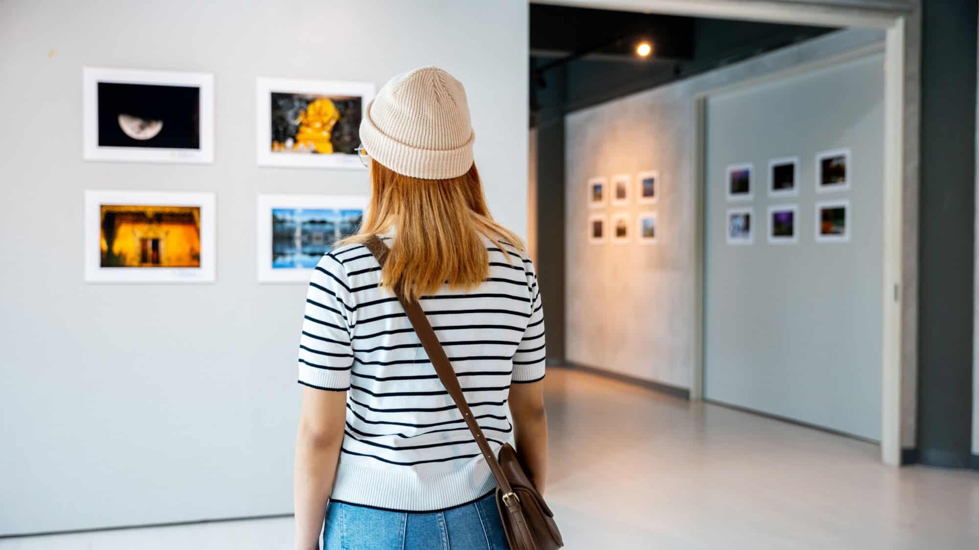 Young woman standing looking art gallery in front of colorful framed paintings pictures on white wall, female watch at photo frame to leaning against at exhibit museum, Back view