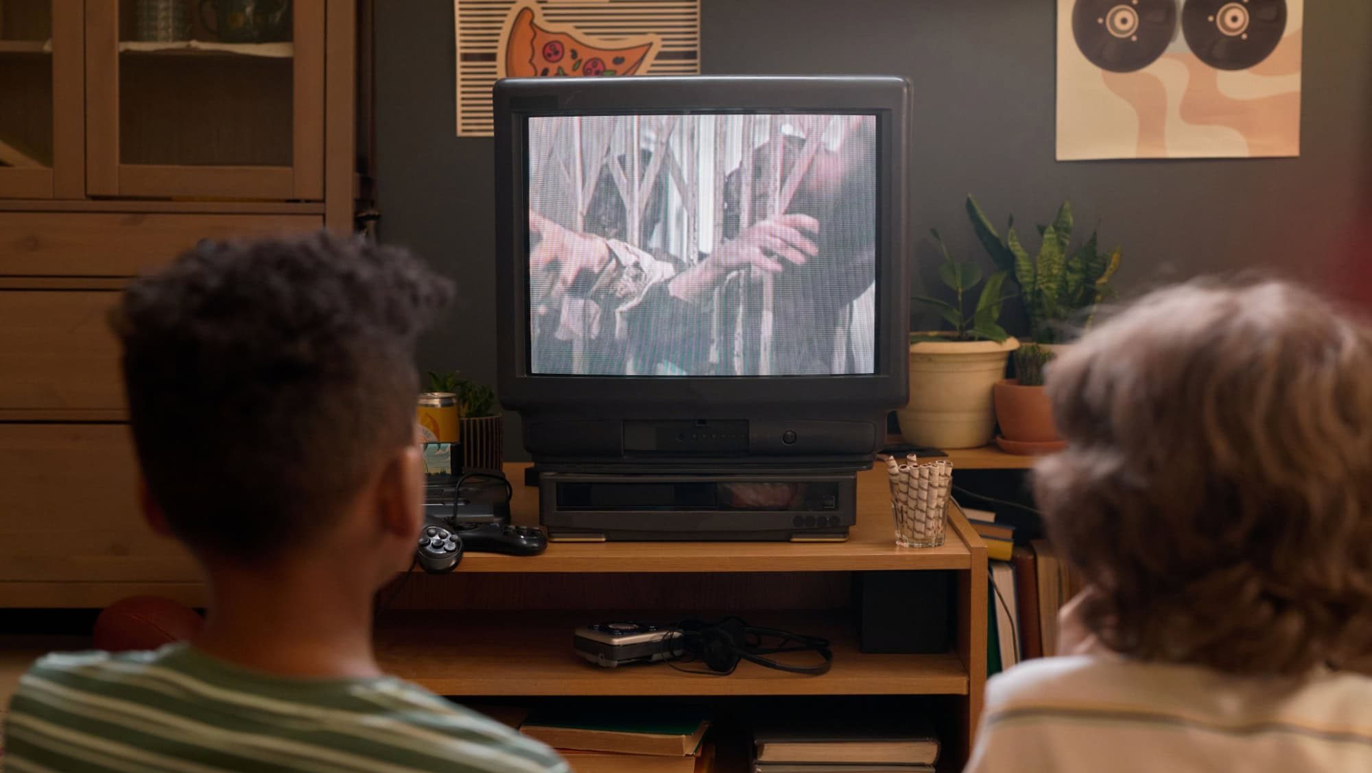 Back view of two kids sitting on floor watching TV showing horror movie about zombies in cozy retro living room with plants and posters on wall, and old-fashioned gaming console below TV screen