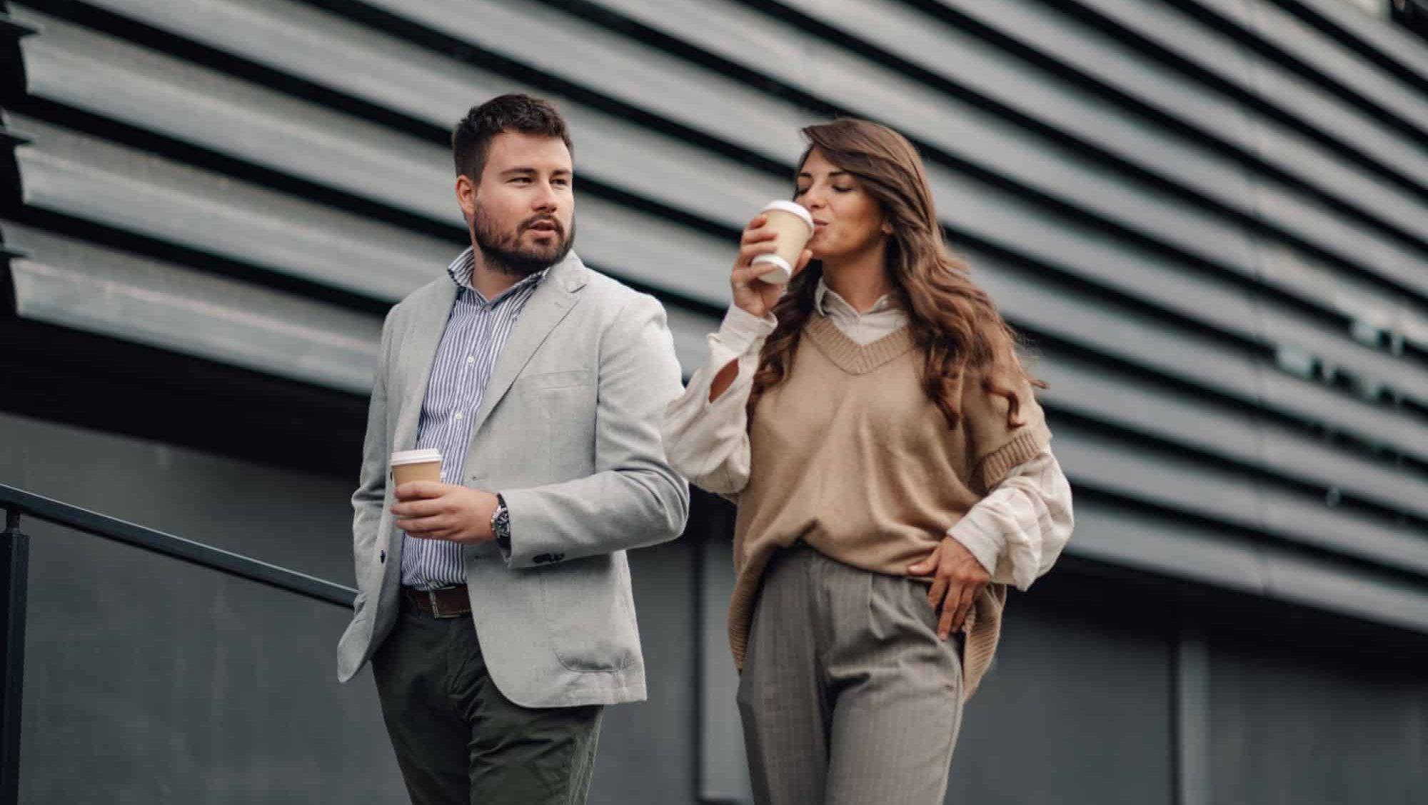 Two colleagues are walking outside the office with takeaway coffee cups, taking a break and having a friendly conversation