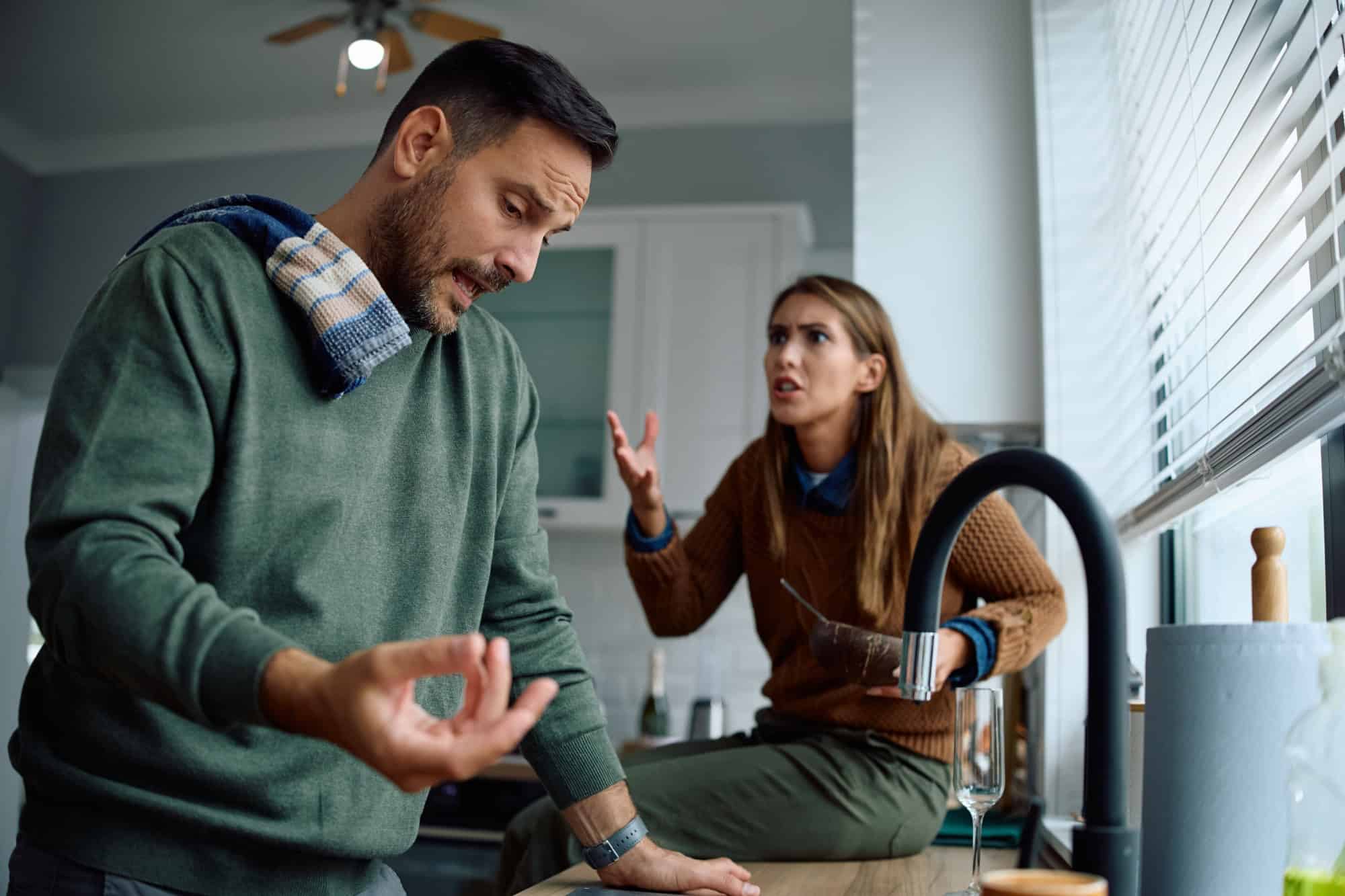 Displeased couple having an argument while washing dishes in the kitchen. Focus is on man.