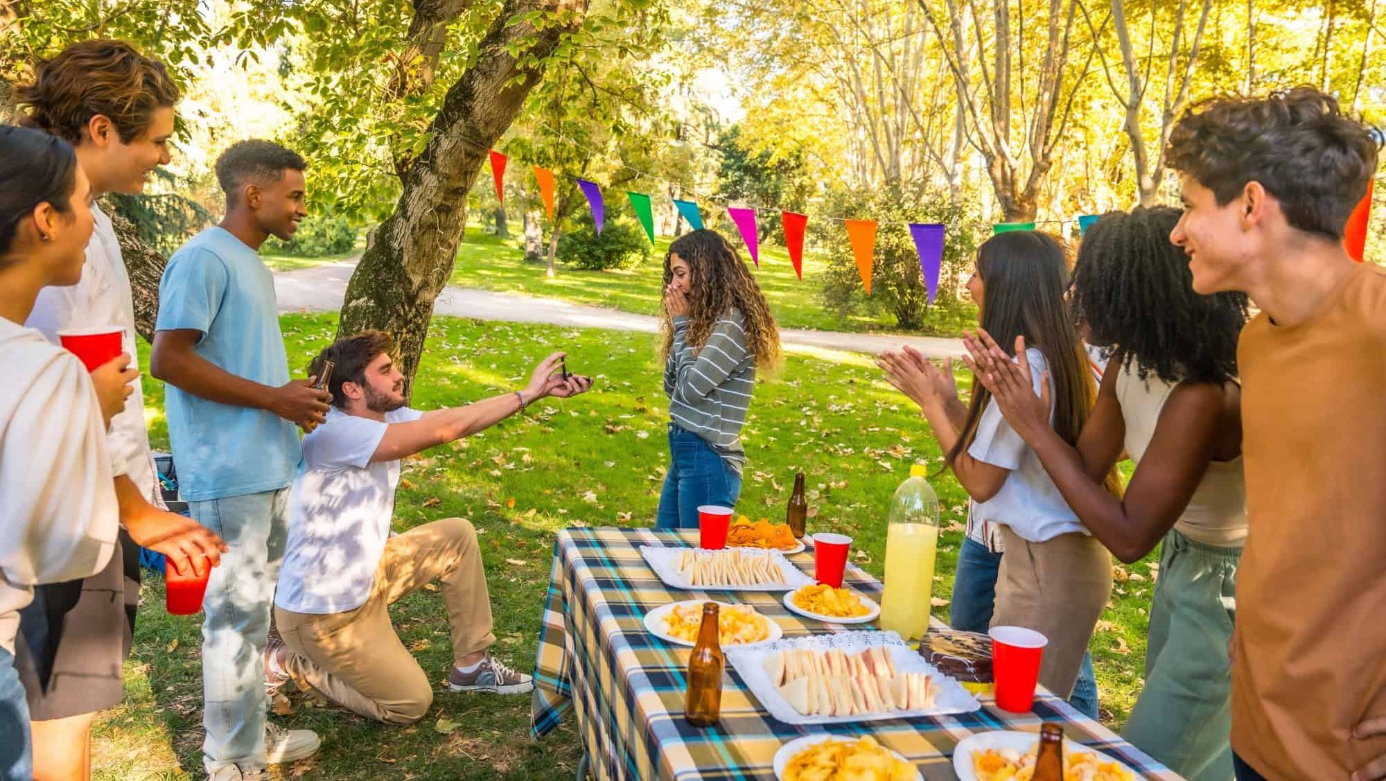 Caucasian young man kneeling to propose marriage to woman during picnic with multi-ethnic friends