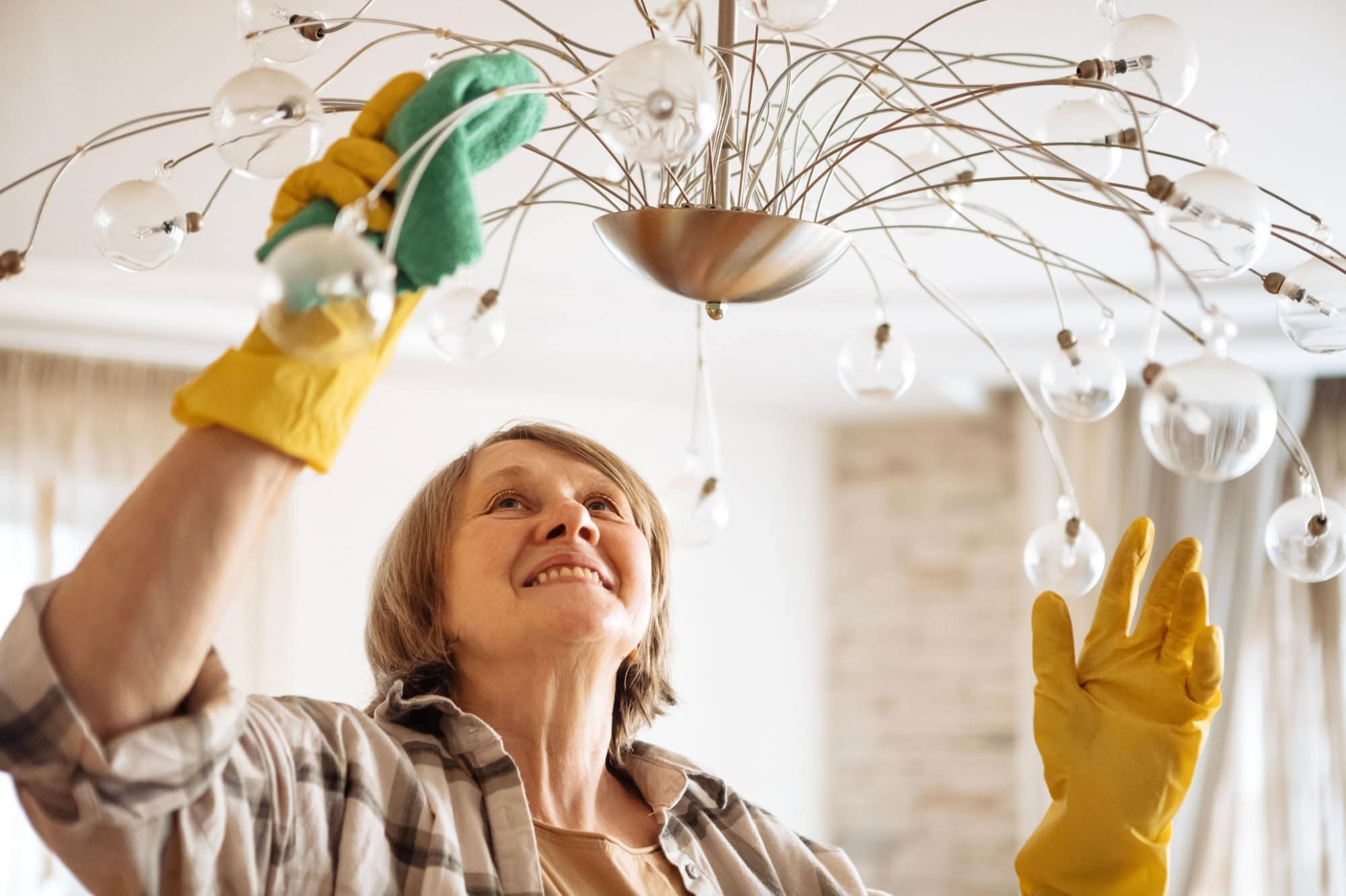 An elderly woman in casual clothes washes a chandelier in the living room. A mature lady with glasses is cleaning the house. A pensioner in rubber gloves smiles and cleans a lamp