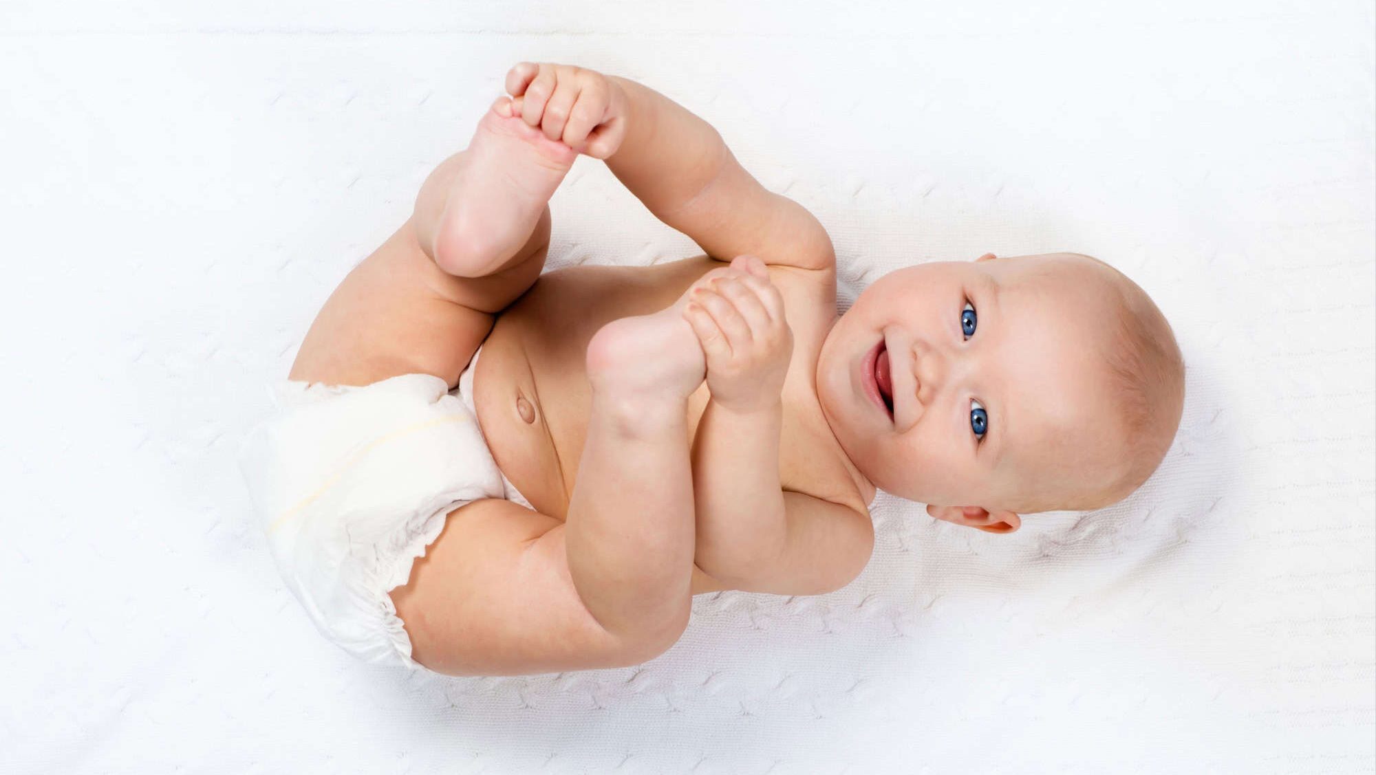 Funny little baby wearing a diaper playing on a white knitted blanket in a sunny nursery. Child after bath or shower on a fresh towel. Infant nappy change and skin care. Cute kid playing with his feet