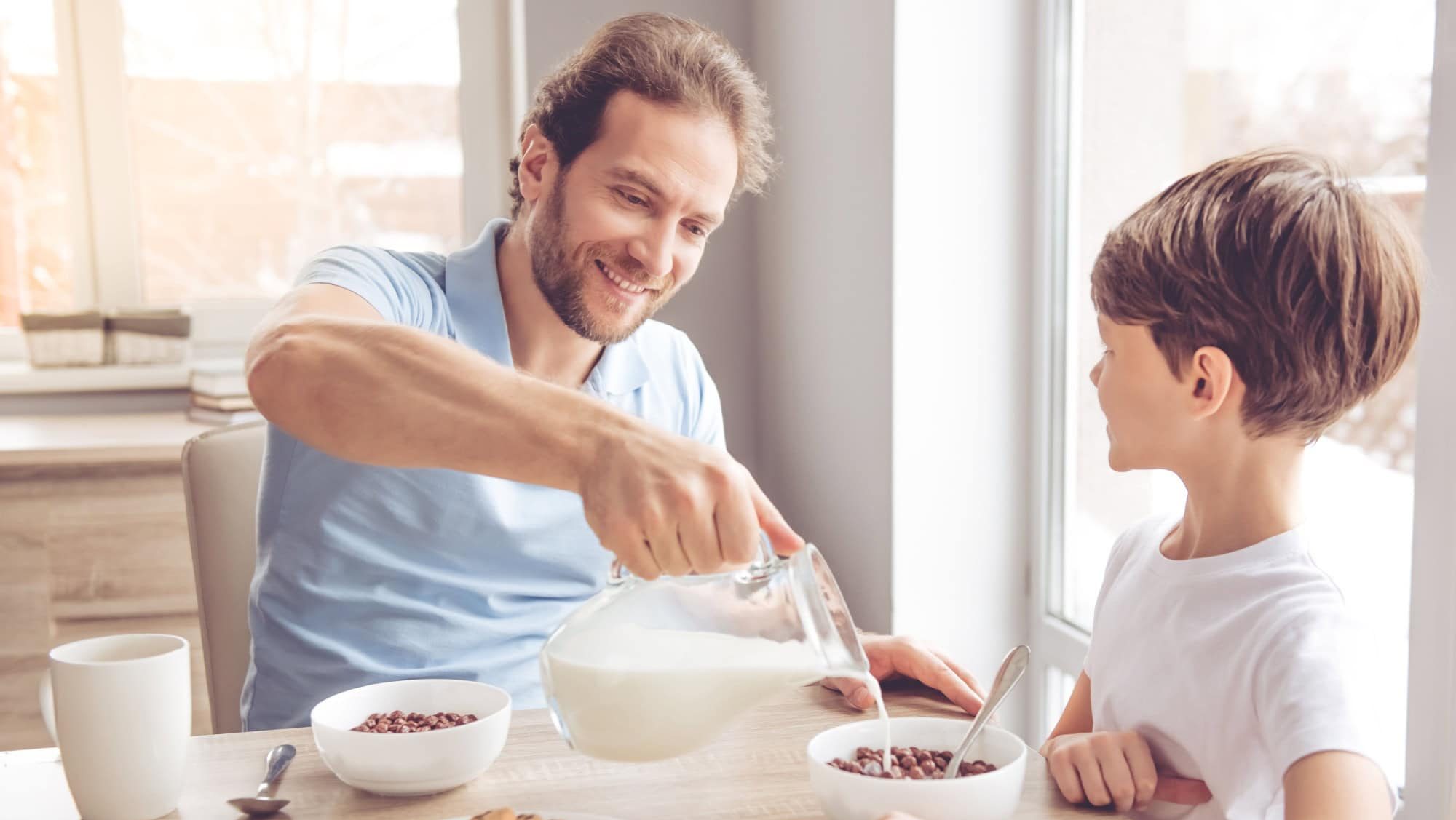 Father and son are smiling while having a breakfast in kitchen. Dad is pouring milk into bowls