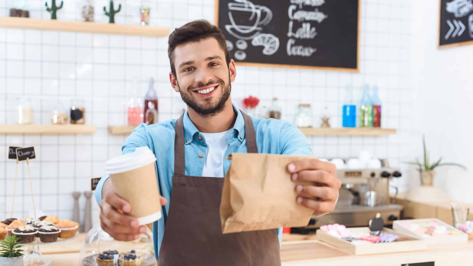 Handsome smiling young barista holding coffee to go in paper cup and take away food in paper bag