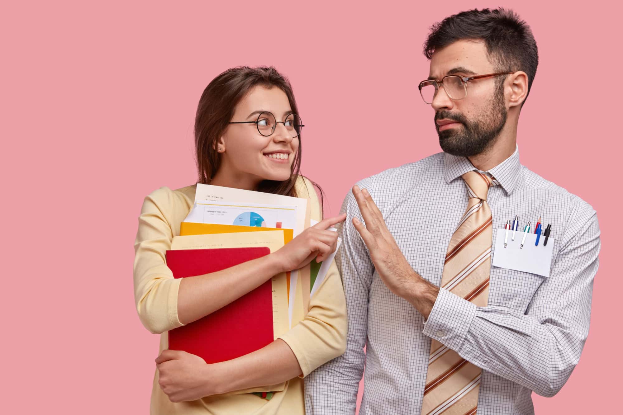 Positive schoolgirl carries papers and notepad, asks friend for help, has glad expression, wears spectacles, isolated over pink background. Bearded young man demonstrates refusal gesture to girlfriend