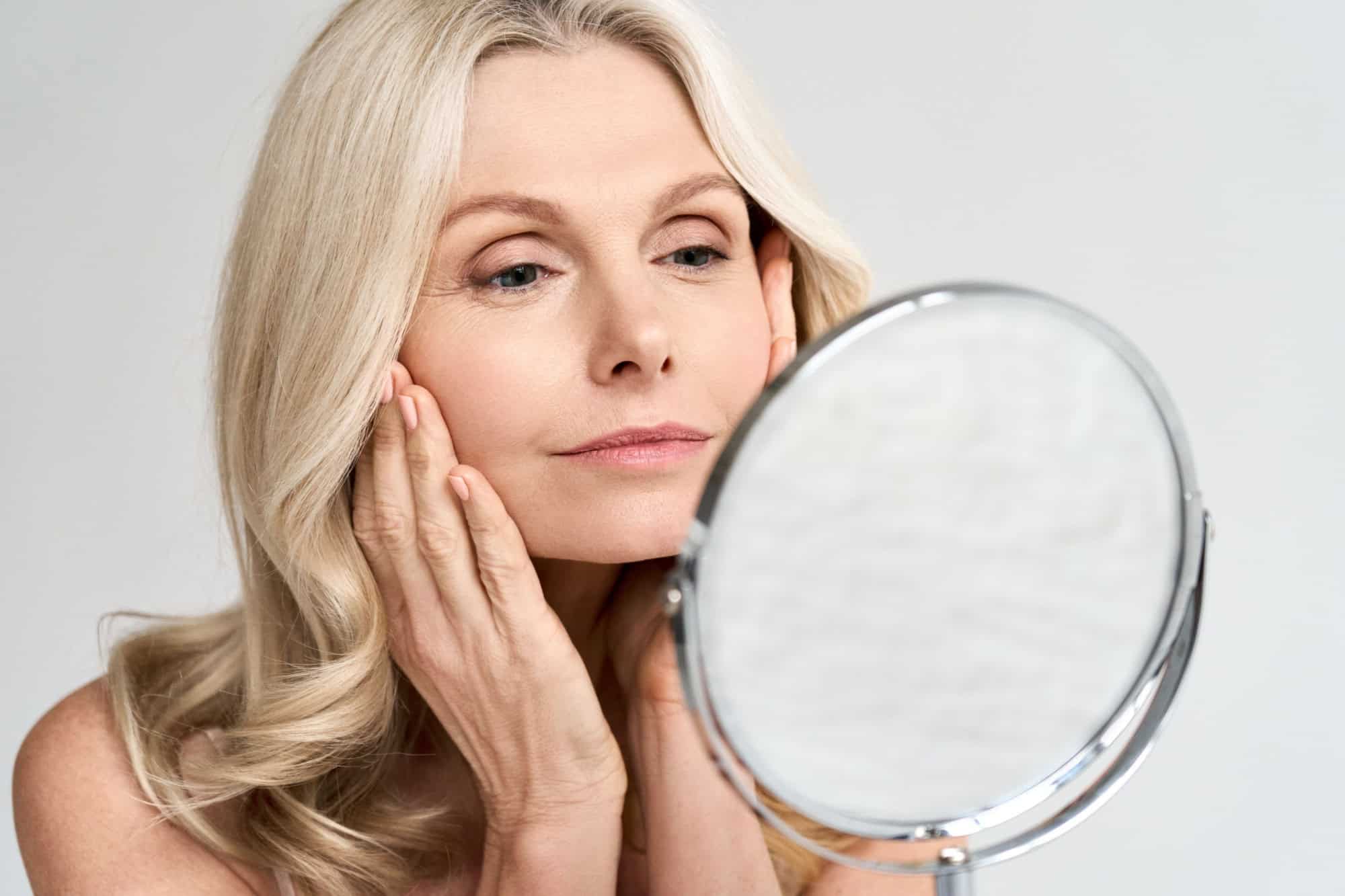Closeup portrait of gorgeous happy middle age woman looking at mirror touching her skin enjoying treatment for dry skin. Advertising of antiaging beauty skin care products.