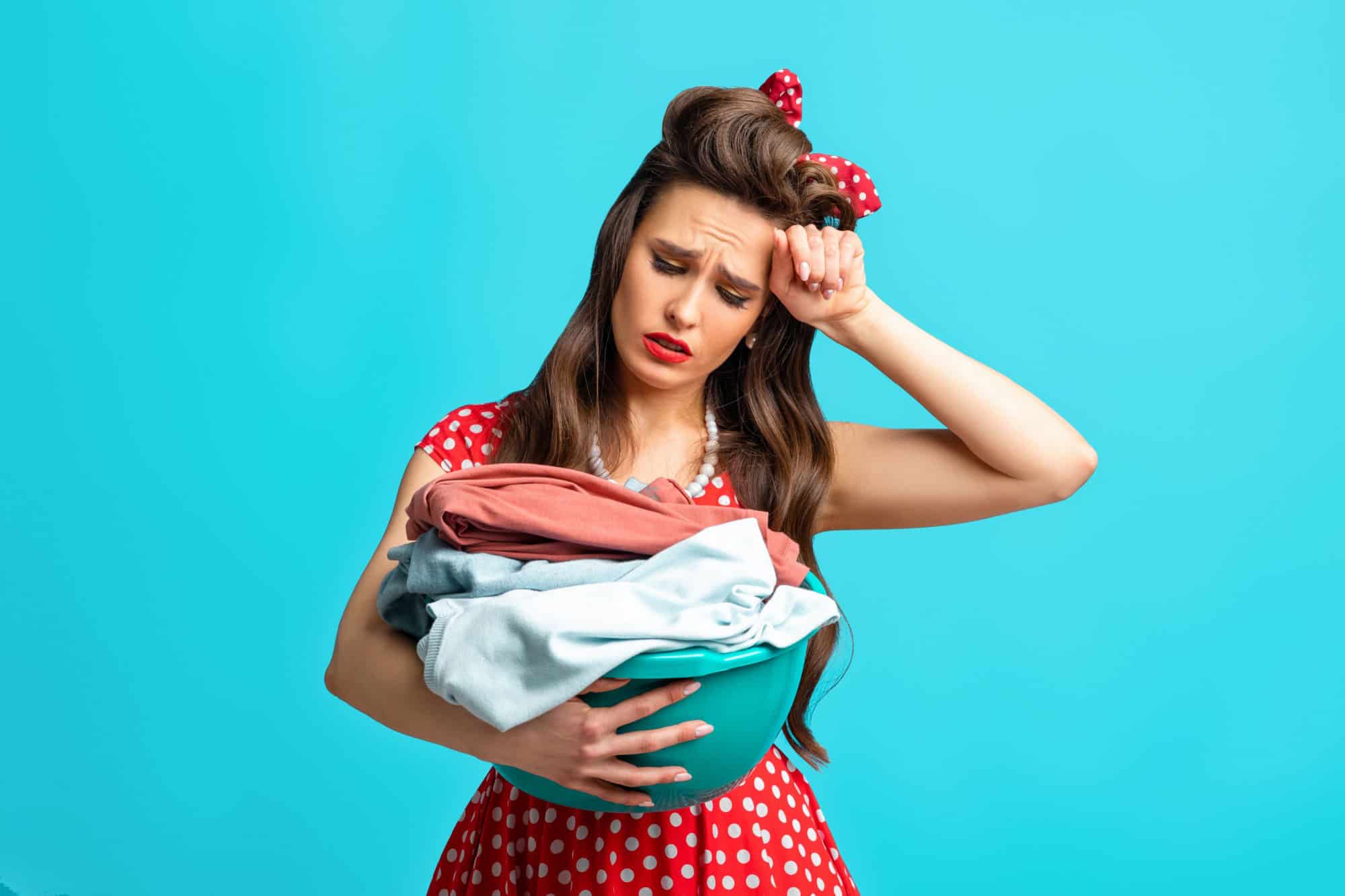 Sad exhausted pinup woman in retro outfit holding clothes for washing or ironing on blue studio background. Millennial housewife in old fashioned dress overwhelmed with too much laundry