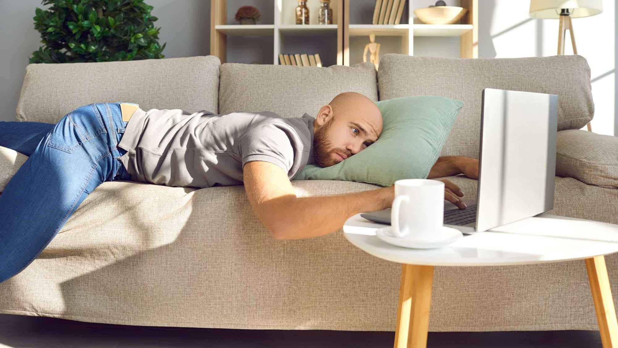Man lying on sofa at home. Sad bored lazy passive guy resting alone on couch, looking at laptop computer screen, missing girlfriend in lockdown, waiting for job recruitment email, thinking about life