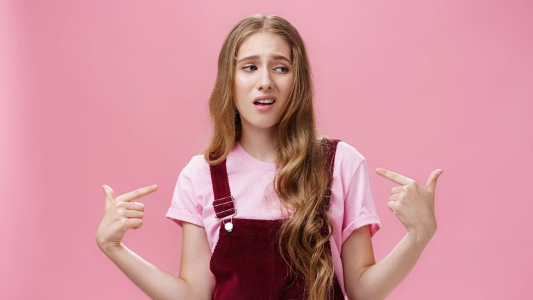 Self-assured arrogant young female student with high ego standing cocky and brag about herself pointing at her with cool snobbish look looking away to right with contempt posing over pink wall