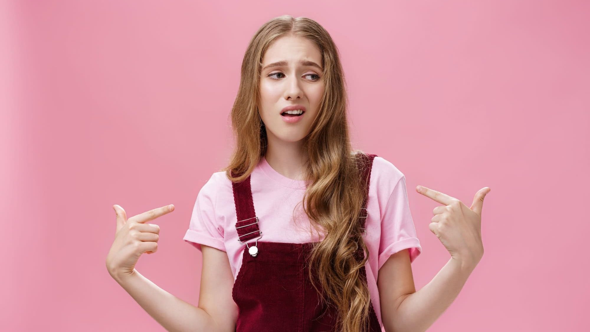 Self-assured arrogant young female student with high ego standing cocky and brag about herself pointing at her with cool snobbish look looking away to right with contempt posing over pink wall