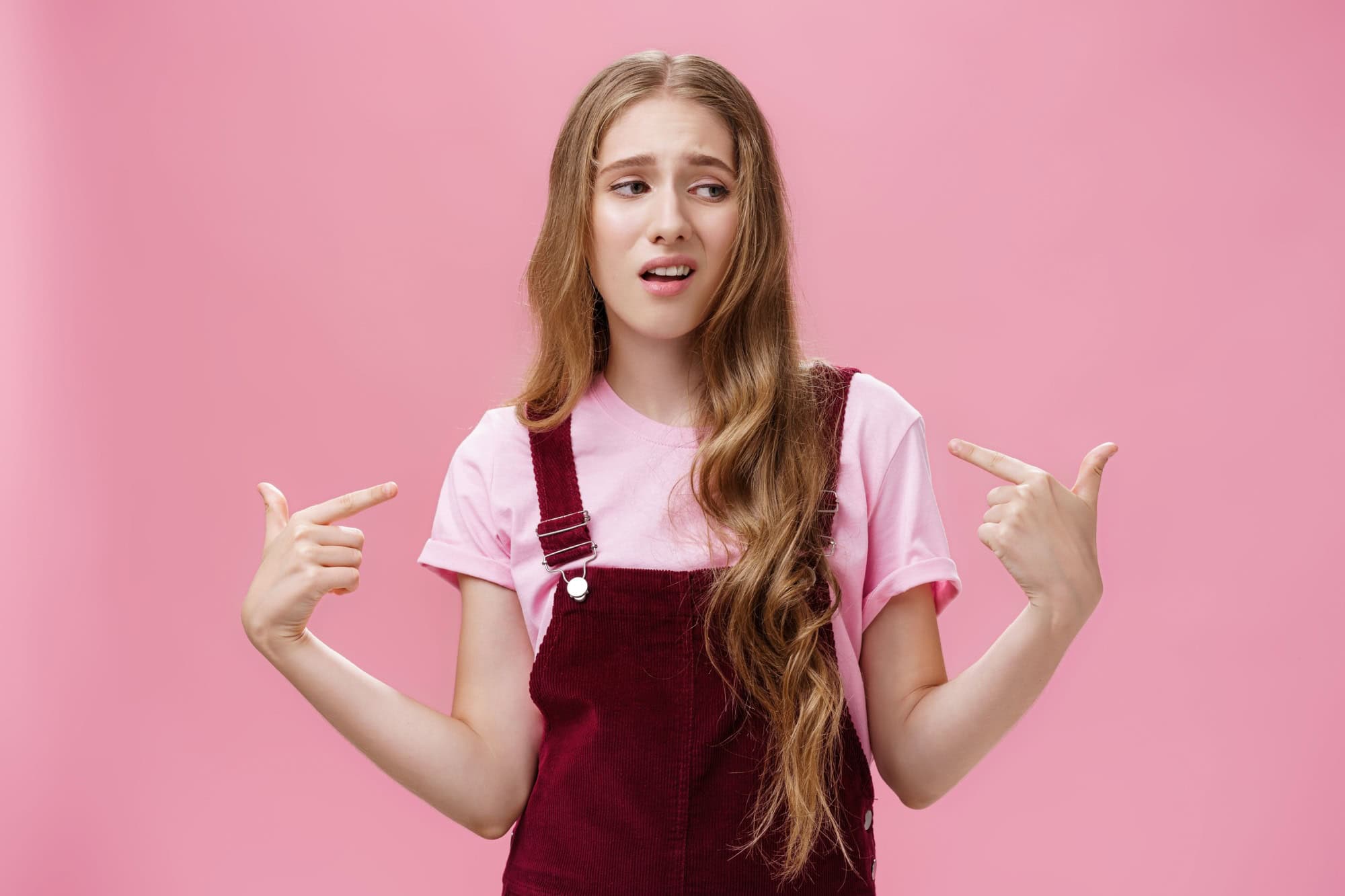 Self-assured arrogant young female student with high ego standing cocky and brag about herself pointing at her with cool snobbish look looking away to right with contempt posing over pink wall