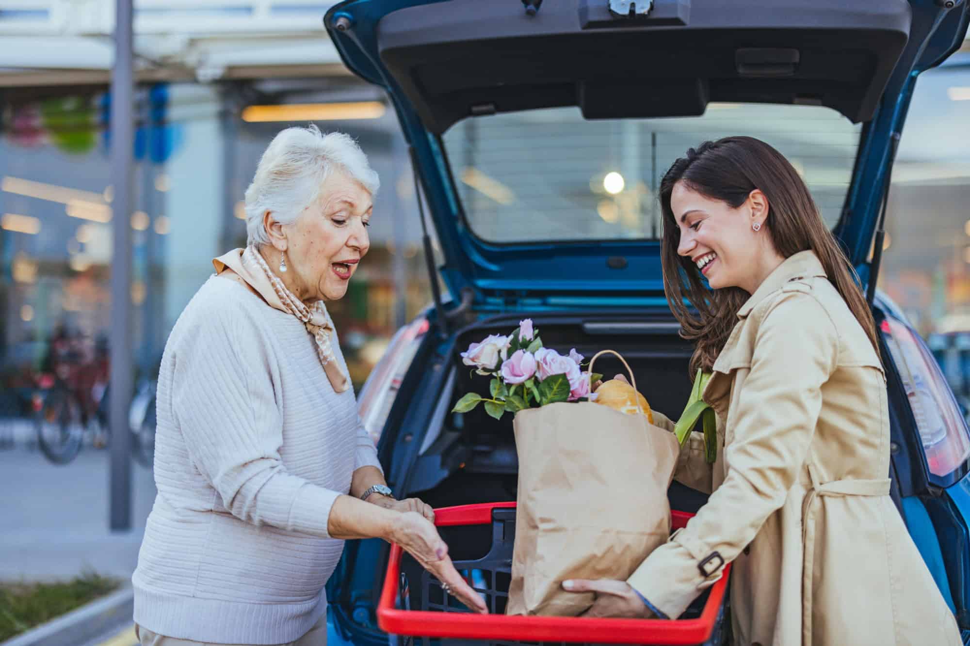 A cheerful Caucasian woman helps her elderly mother with grocery bags, placing them into the car trunk in a parking lot, reflecting a caring family moment.