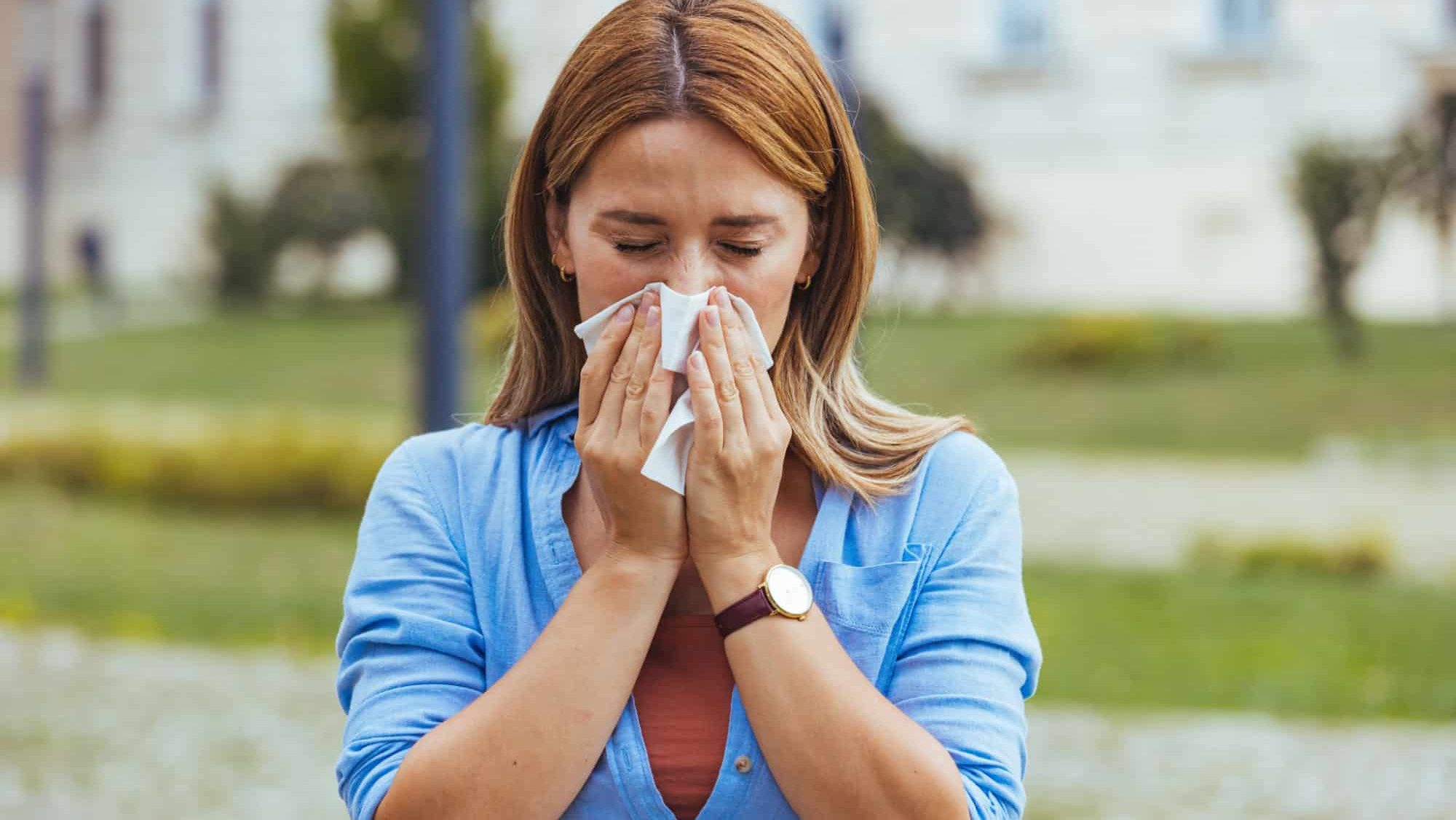A young woman sneezes into a tissue while standing outside, suggesting allergies or a cold. The outdoor setting emphasizes natural triggers and health concerns.
