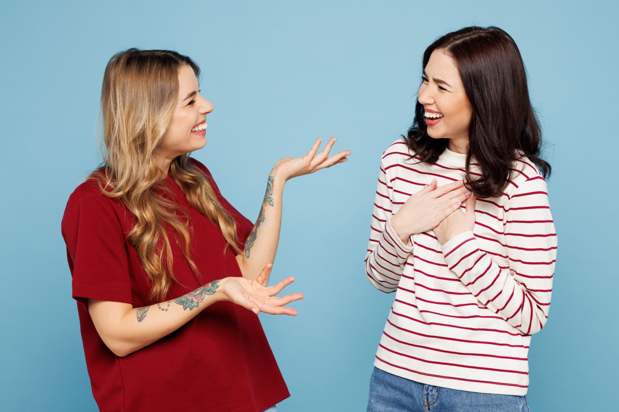 Side view young couple two friends women wearing red casual clothes together talk speak to each other tell joke isolated on pastel plain light blue cyan background studio portrait. Lifestyle concept