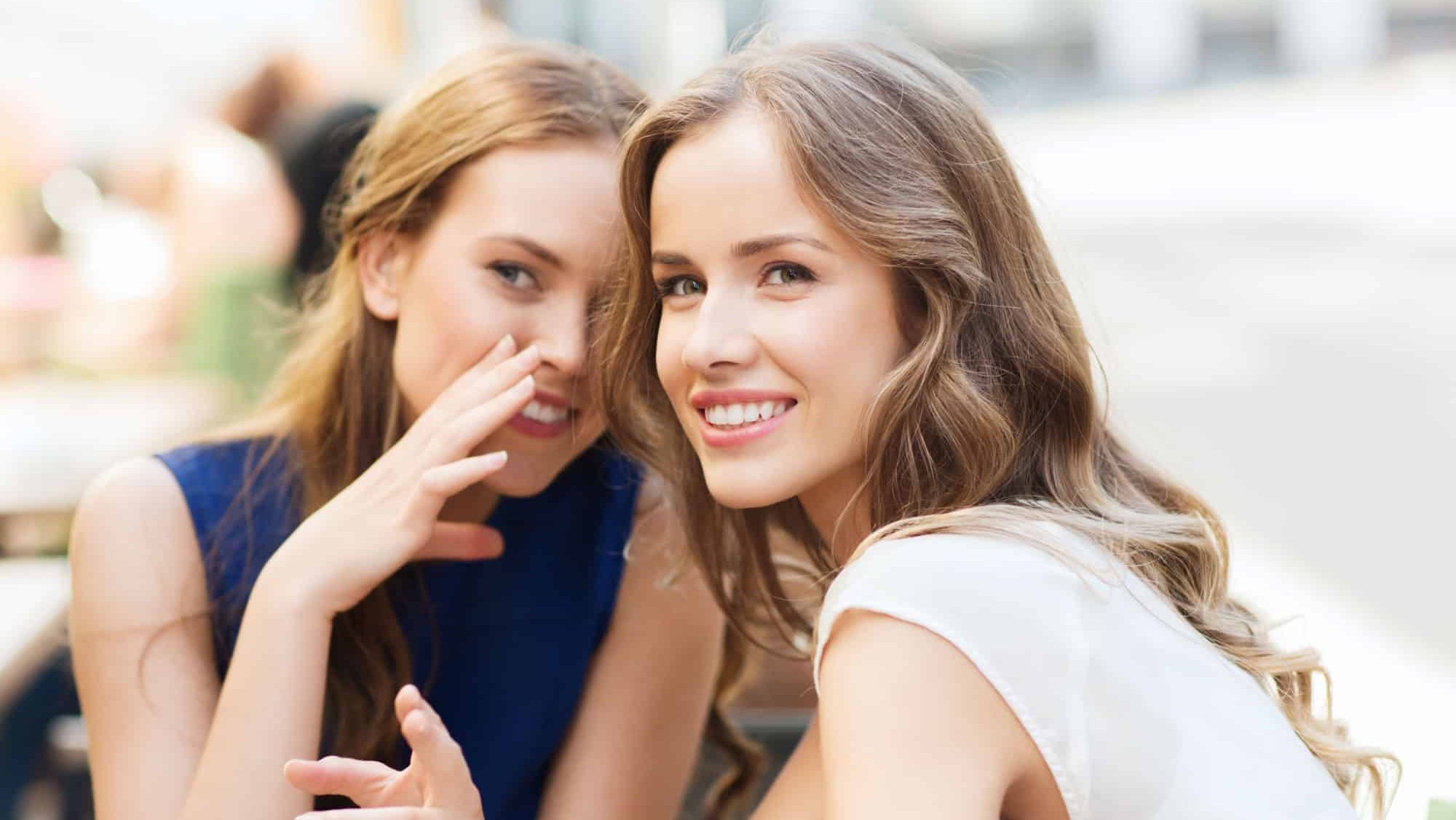 People communication and friendship concept - smiling young women drinking coffee or tea and gossiping at outdoor cafe
