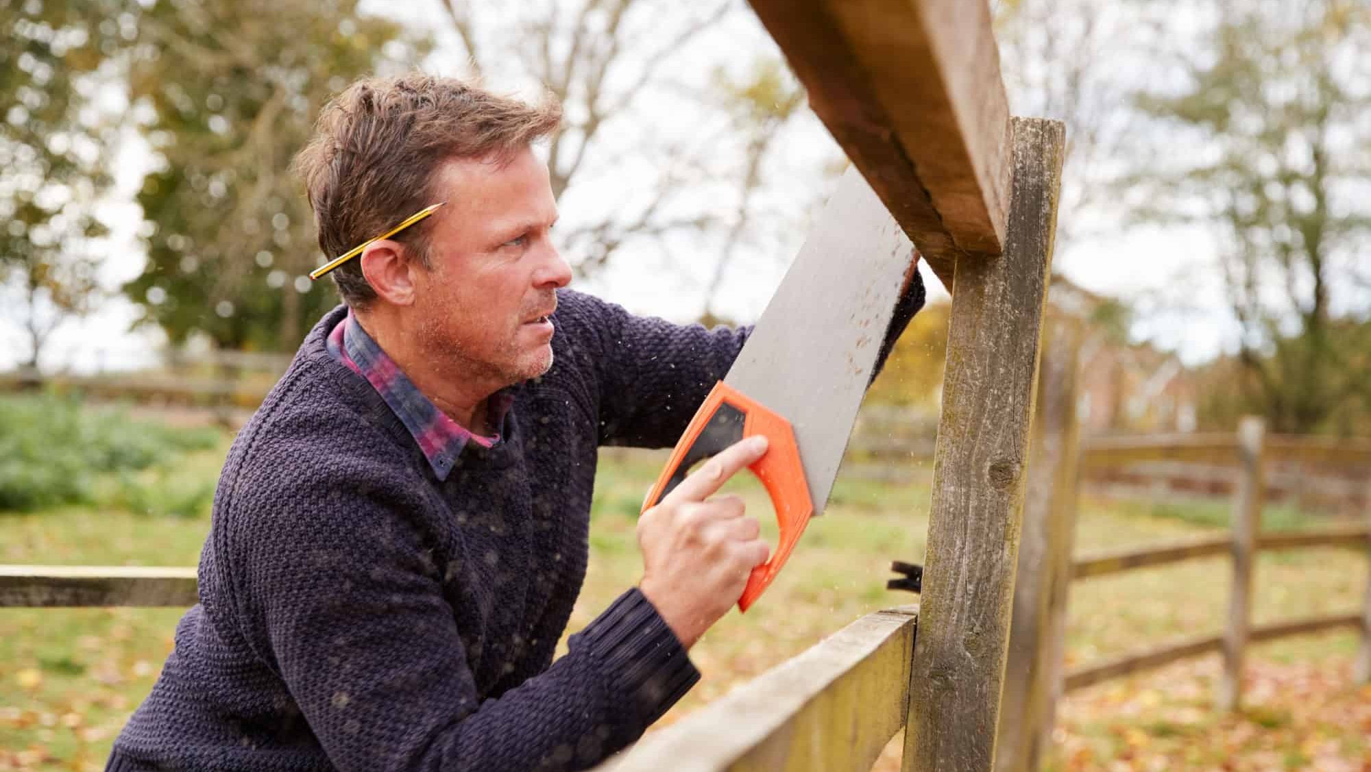 Mature Man Fixing Outdoor Fence With Saw