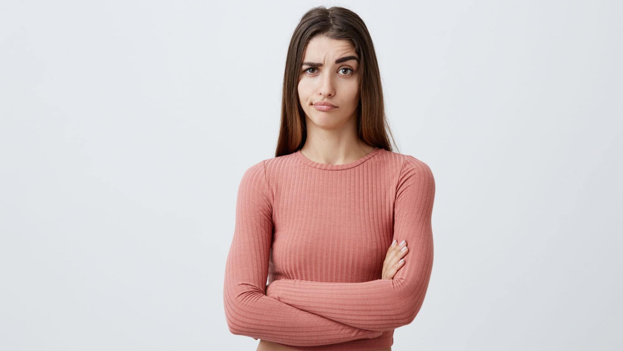 Beautiful young unhappy Caucasian brunette girl with long hair in fashionable outfit crossing hands, looking in camera with sarcastic expression, can't believe her boyfriend was with friend last night