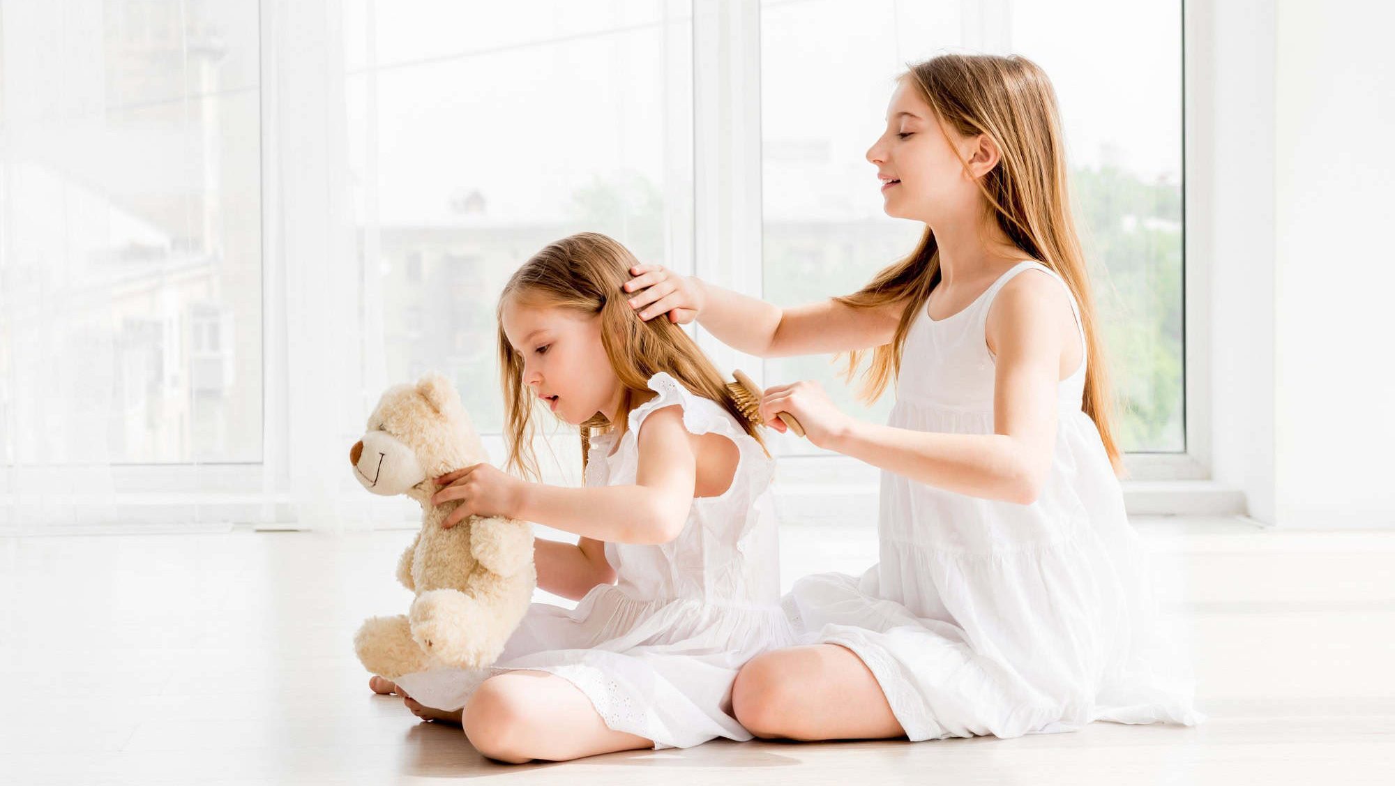 Lovely little girl brushing hair of her younger sister while sitting on the floor. Little child girl playing with bear toy while her older sister combing her hair