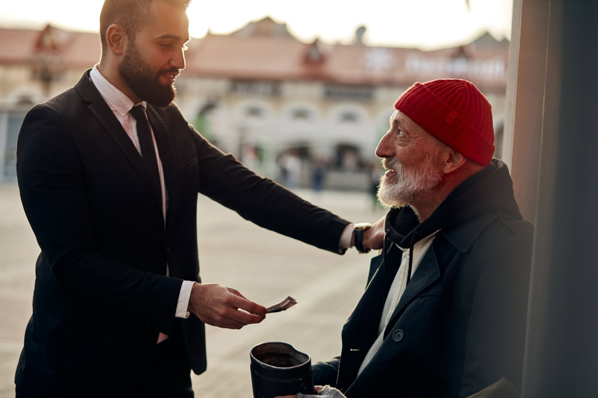 Businessman in tuxedo help to vagrant man sitting on street with rusty money jar. Rich man put one hand on shoulder of homeless person and give one dollar. Kind people, compassion, sympathy