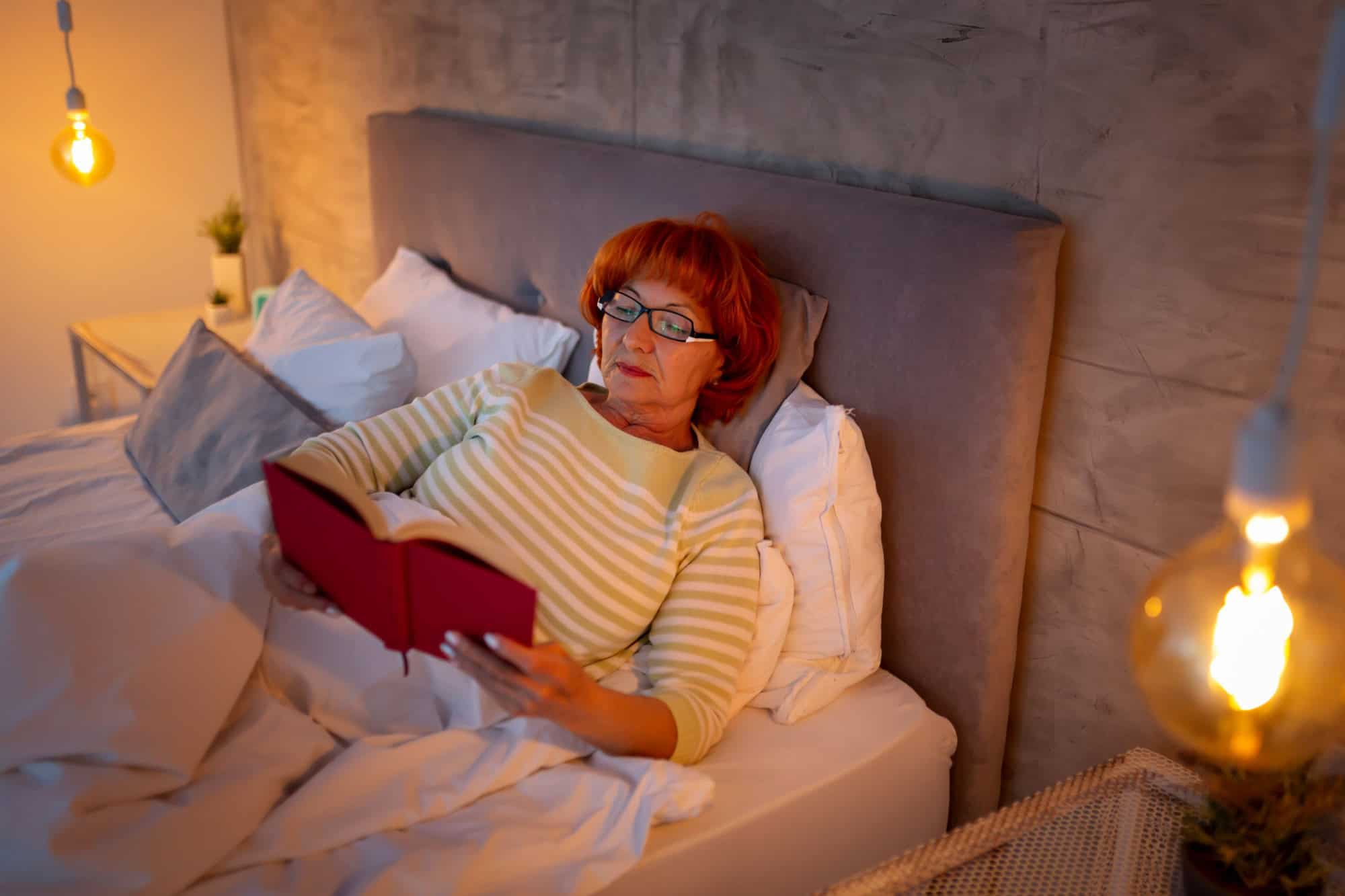Elderly woman wearing pajamas lying in bed, relaxing at home in the evening, reading a book