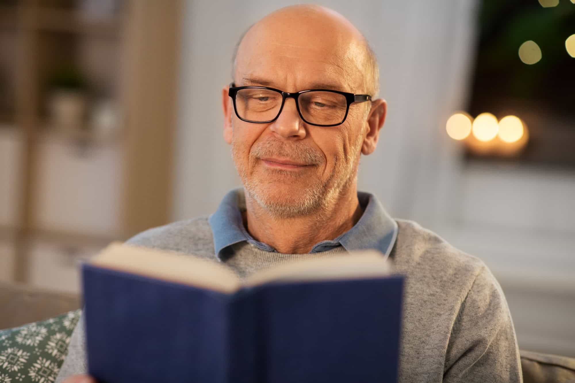 old age, leisure and people concept - happy bald senior man sitting on sofa and reading book at home in evening