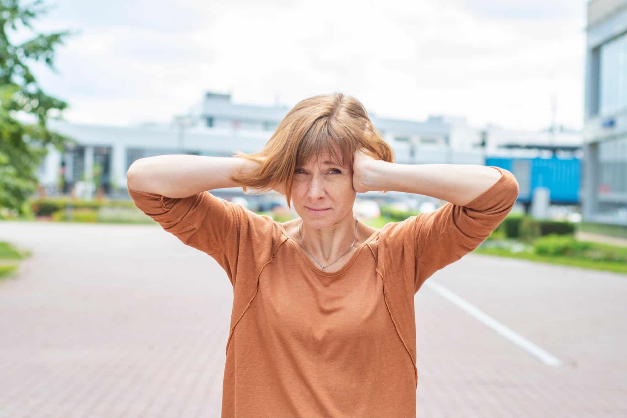 A middle-aged redhead woman in casual clothes stands on the street covering her ears with her hands. Concept - sound pollution of the environment in the city. There are too many loud sounds.