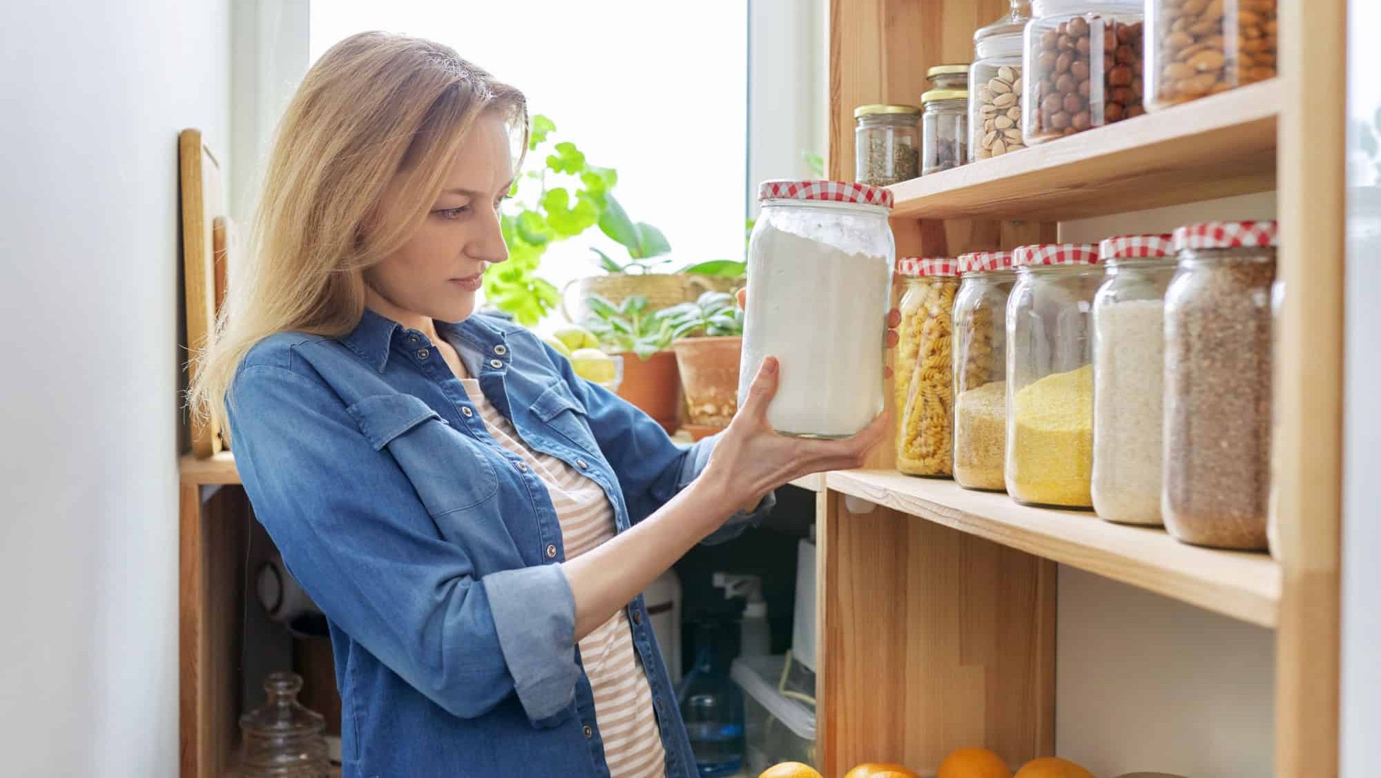 Middle aged woman at home in kitchen in pantry, female looking at food on wooden storage shelves holding jar of flour