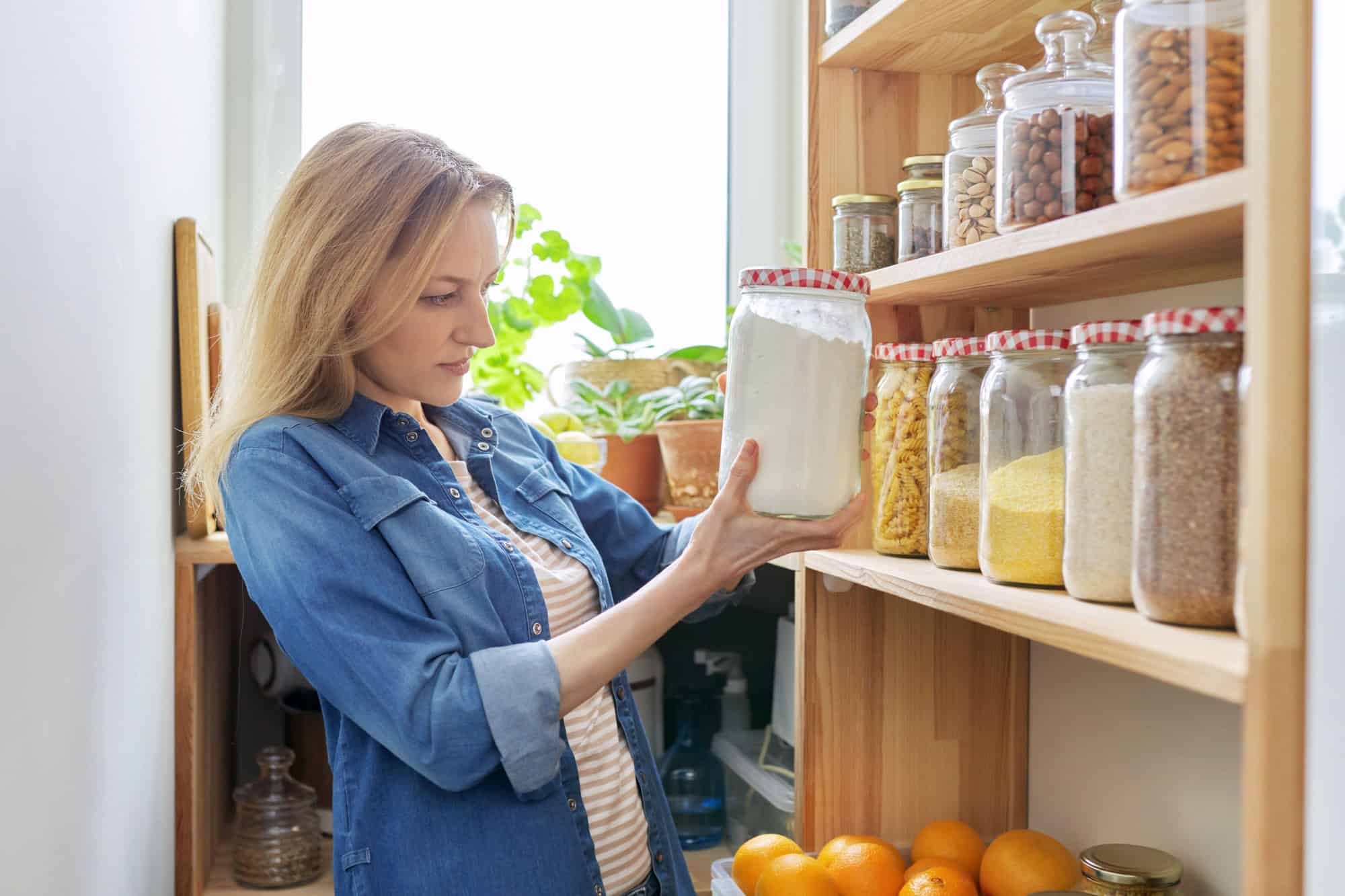 Middle aged woman at home in kitchen in pantry, female looking at food on wooden storage shelves holding jar of flour