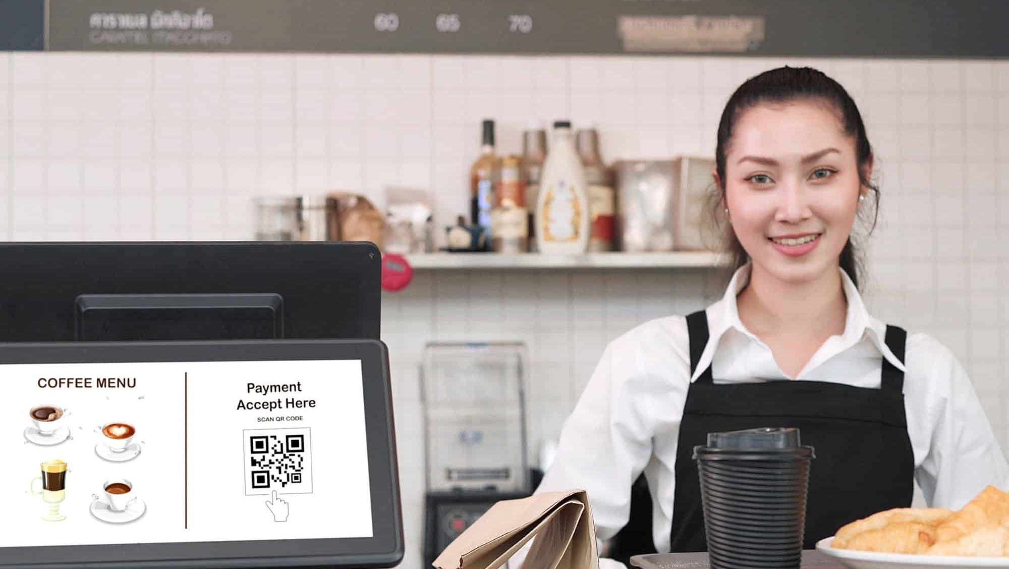 Young Asian woman waitress standing in back counter whlie putting tray with coffee cup and bread, croissants on counter in coffee shop. Menu and QR code payment in screen monitor on counter