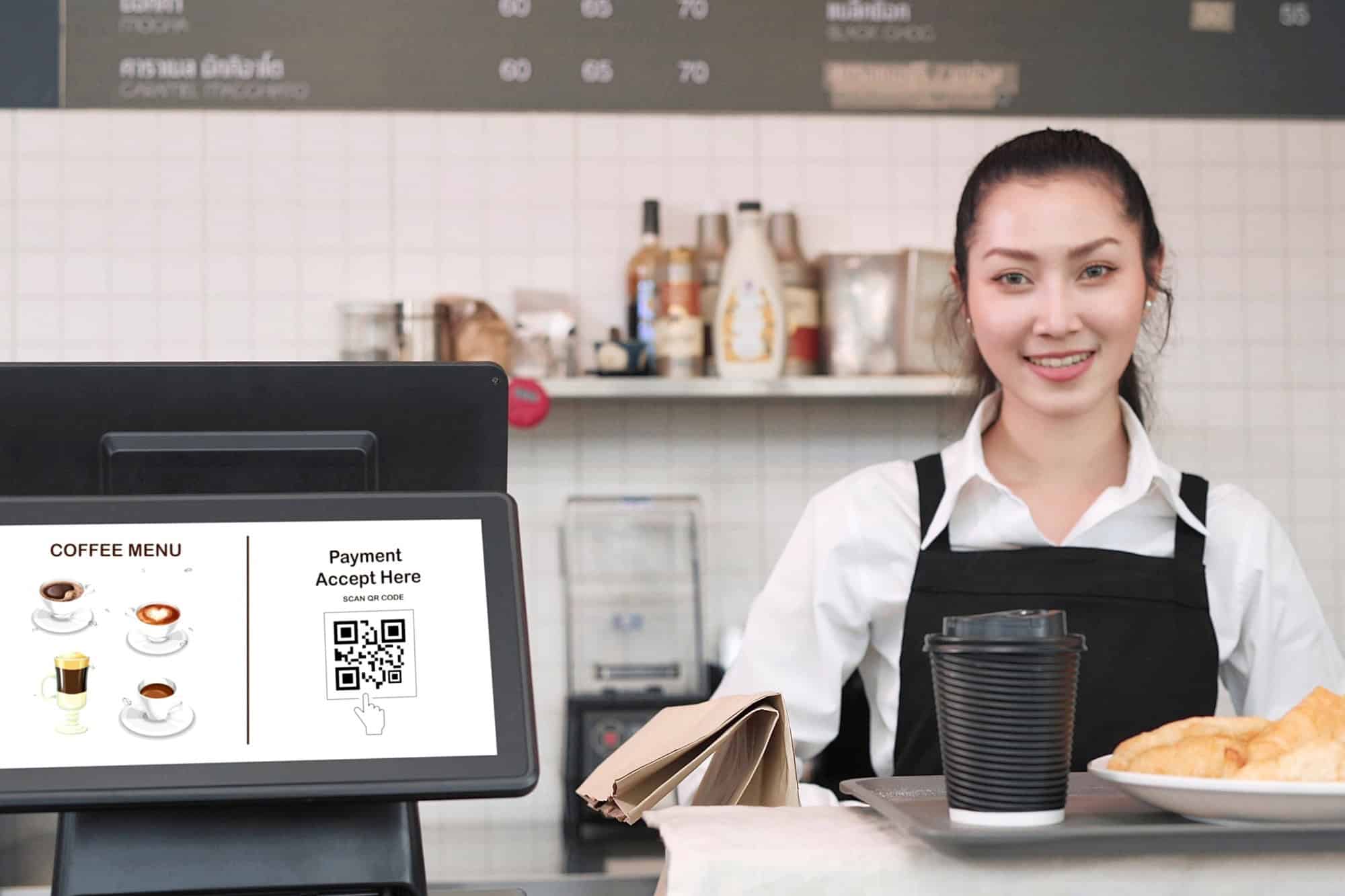 Young Asian woman waitress standing in back counter whlie putting tray with coffee cup and bread, croissants on counter in coffee shop. Menu and QR code payment in screen monitor on counter