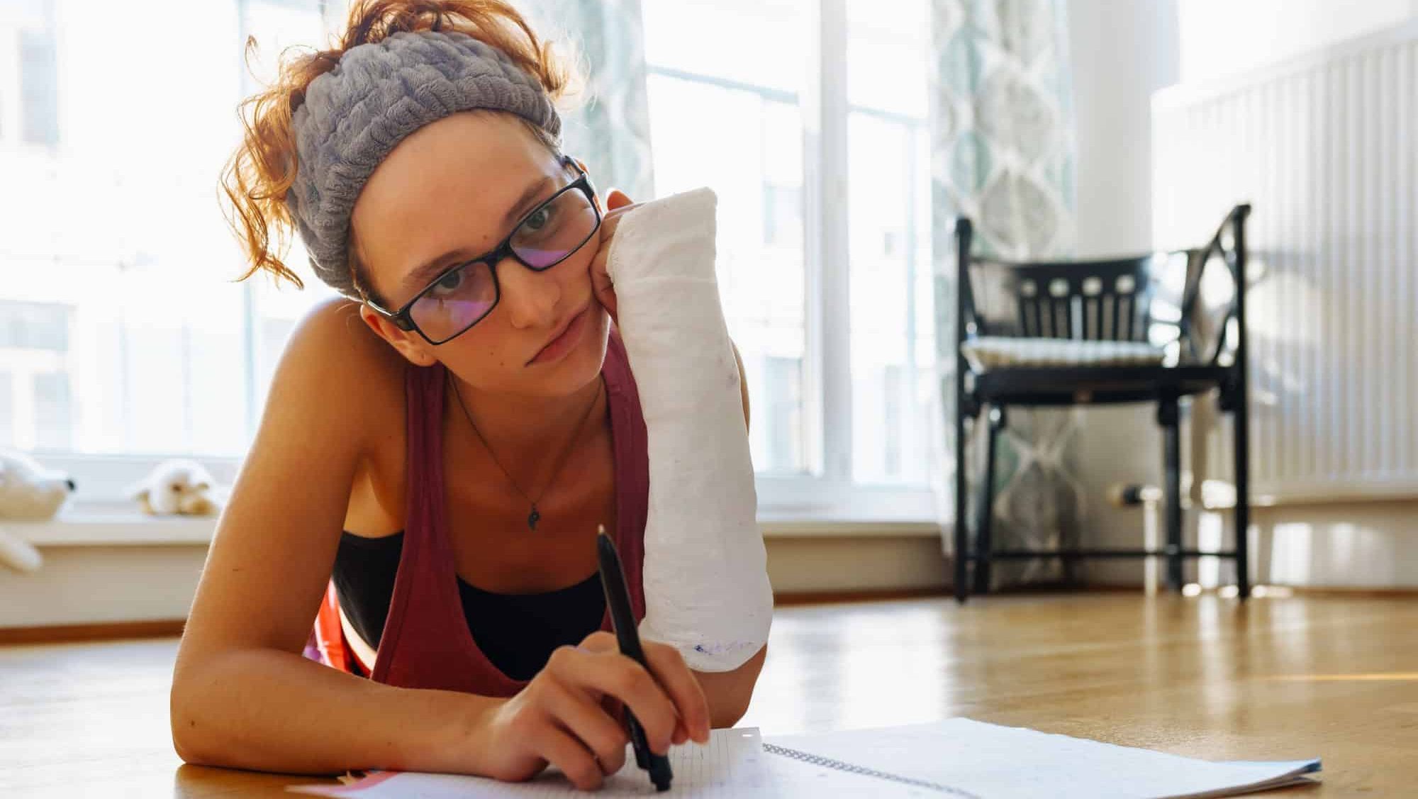 Redheaded teenage girl with broken arm in cast, wearing glasses, lies on parquet floor, resting head on hand, studying, writing notes in notebook, doing homework, at home