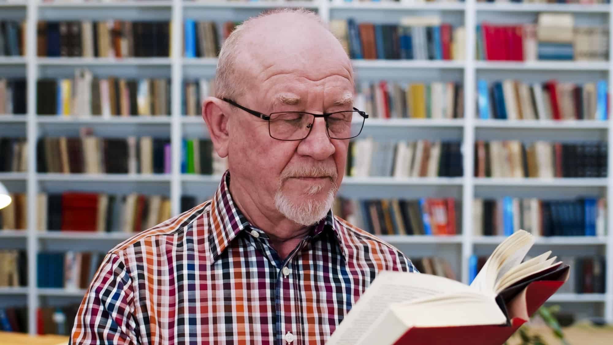 Old Caucasian bald man in glasses and motley shirt reading book in library and studying. Close up of male professor with textbook researching in bibliotheca. Scientific work concept.