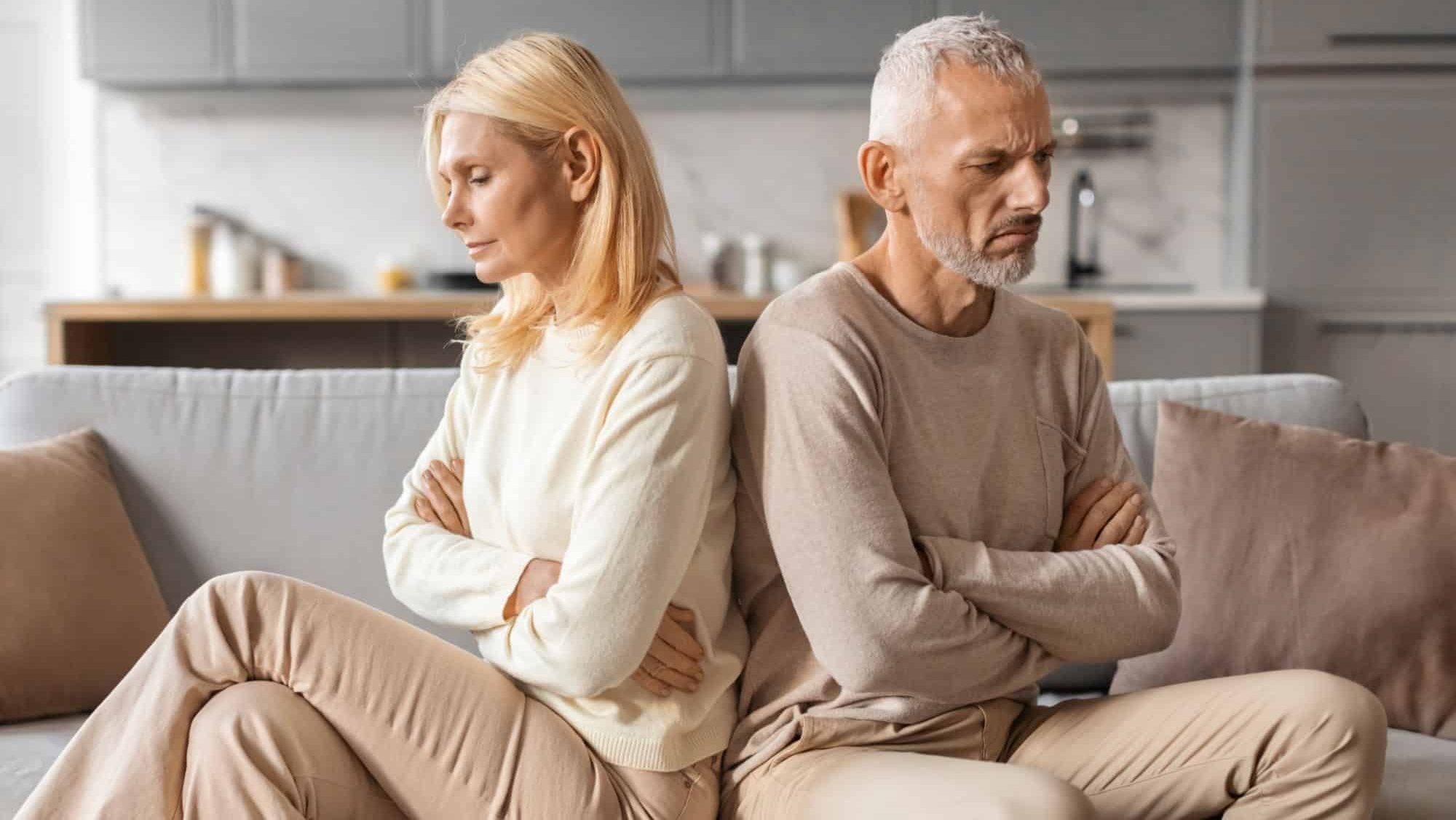 Upset senior married couple grey-haired man and blonde woman sit back to back on couch at home, not talk to each other, feeling down after fight, have difficulties in marriage, thinking about divorce