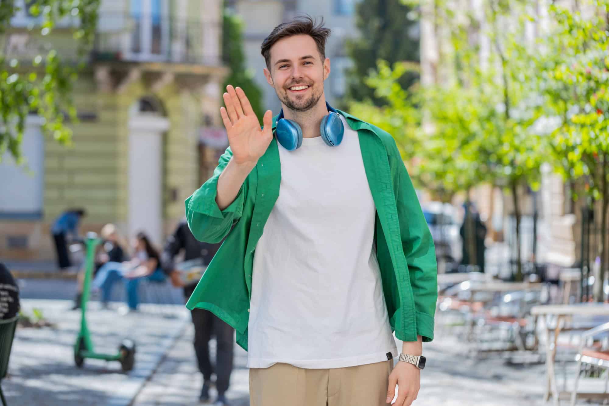Cheerful young man tourist smiling friendly at camera, waving hands gesturing invitation hello, hi, greeting, goodbye, welcoming with hospitable expression outdoors. Guy on urban city town street.