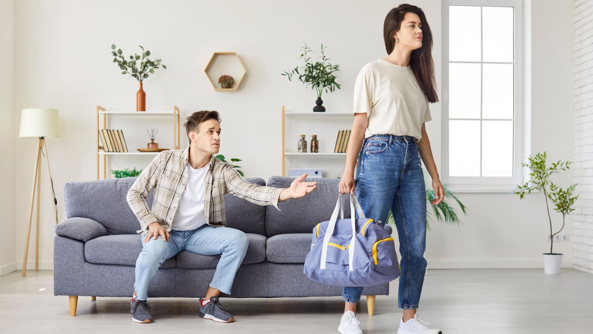 Sad wife with bag walking away from husband during a conflict or divorce at home living room. Negative motions of breakup, bad family relationships, and domestic quarrels into couples.