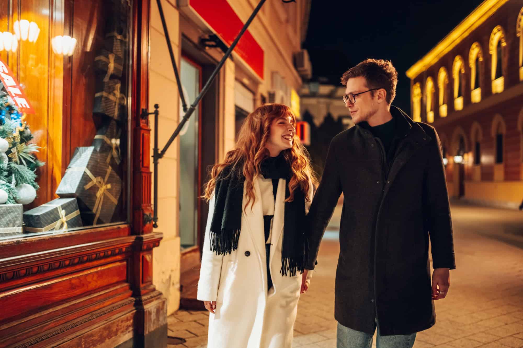 A loving christmas couple is holding hands and walking on city street downtown near store with christmas presents at new year's eve. An affectionate festive couple walking on street on christmas eve.