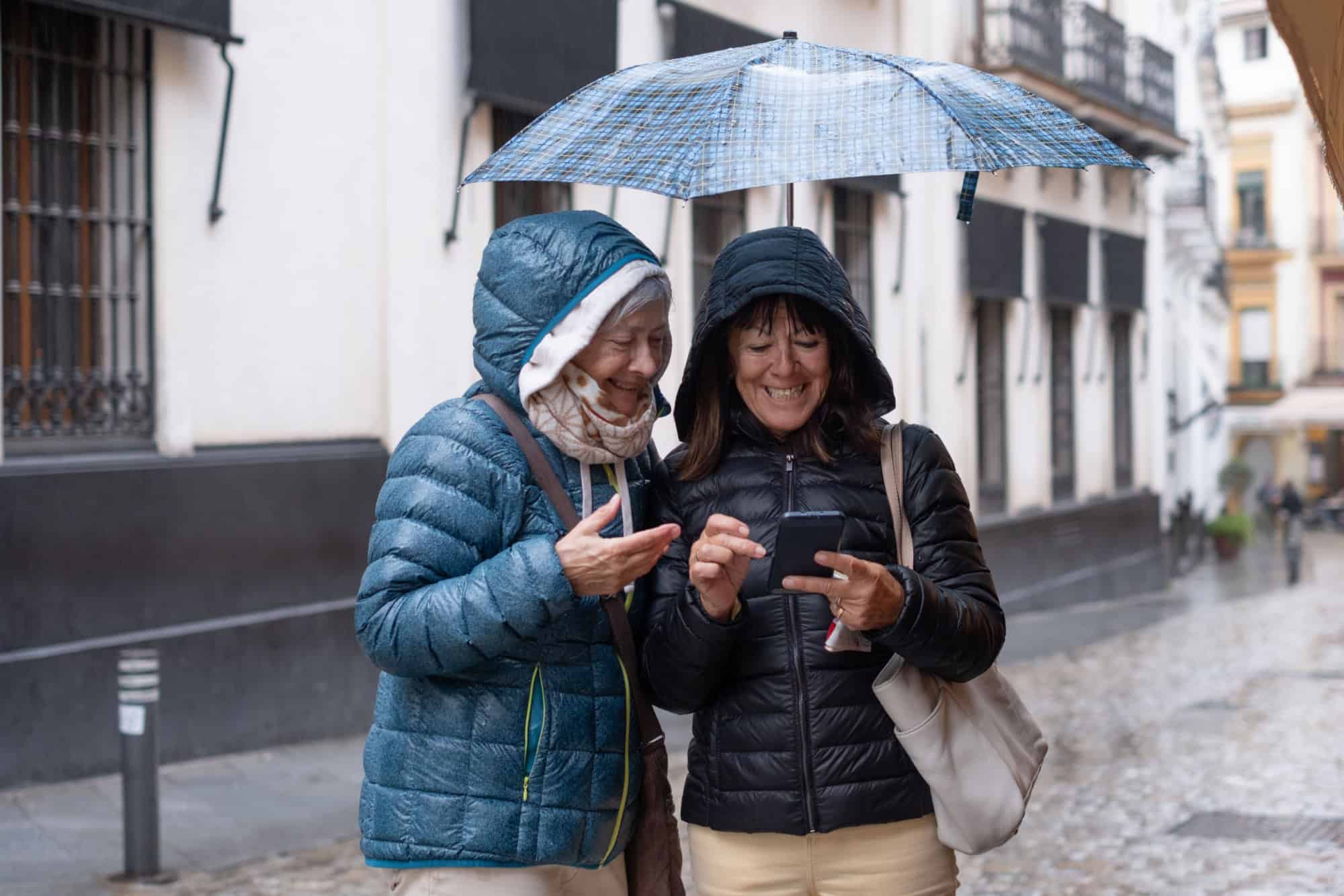 Joyful friendship in the rain. Two elderly women taking sightseeing tour and checking the right direction on their cell phones, standing under an umbrella