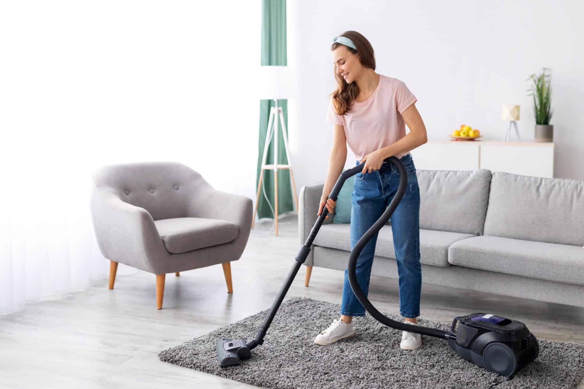 Cheerful female housekeeper using vacuum cleaner to tidy apartment, full length portrait. Domestic hygiene concept