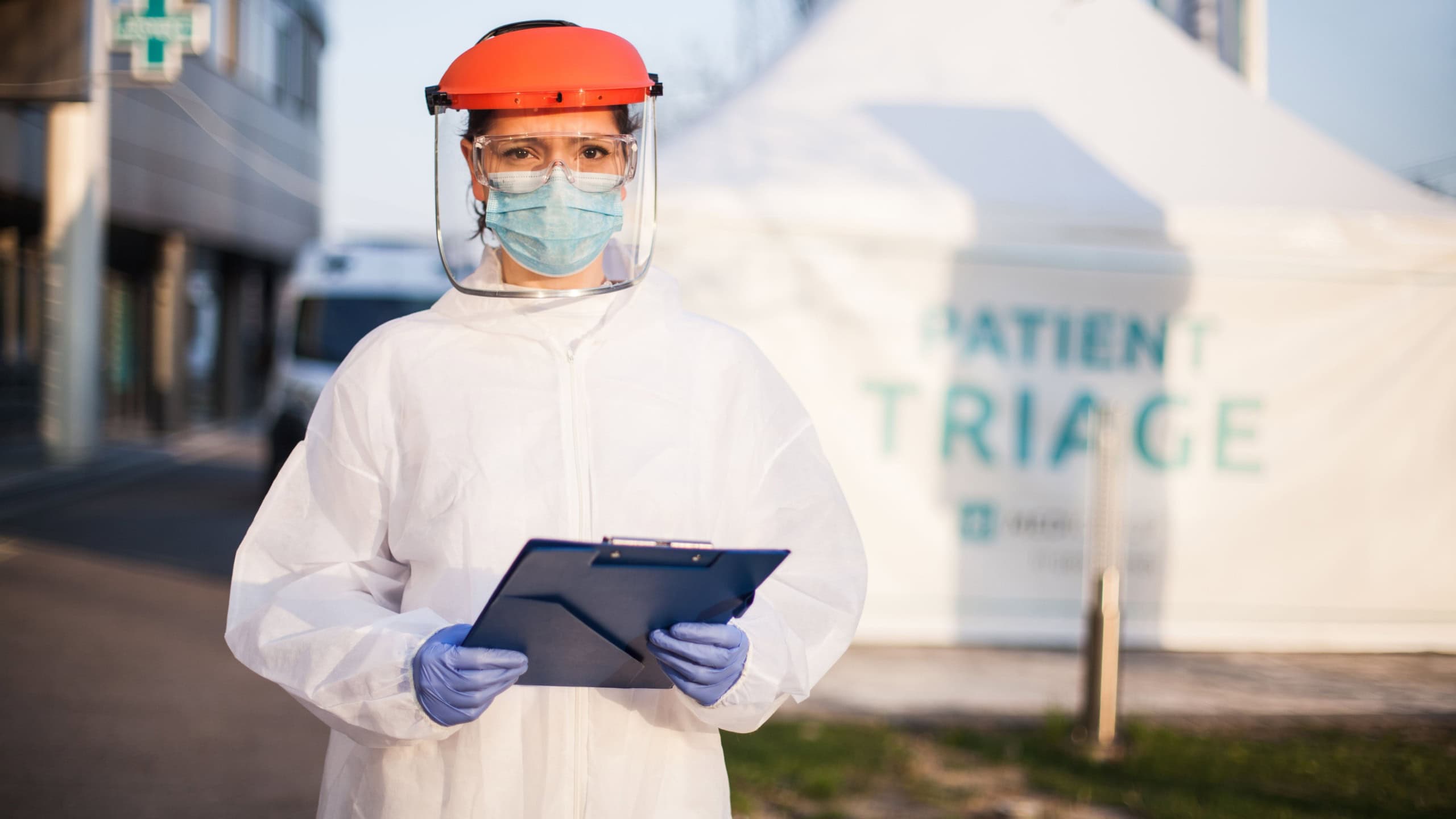 Paramedic wearing personal protective equipment PPE holding folder standing in front of ICU hospital isolation rt-PCR drive thru testing site,COVID-19 pandemic outbreak crisis,worried exhausted staff