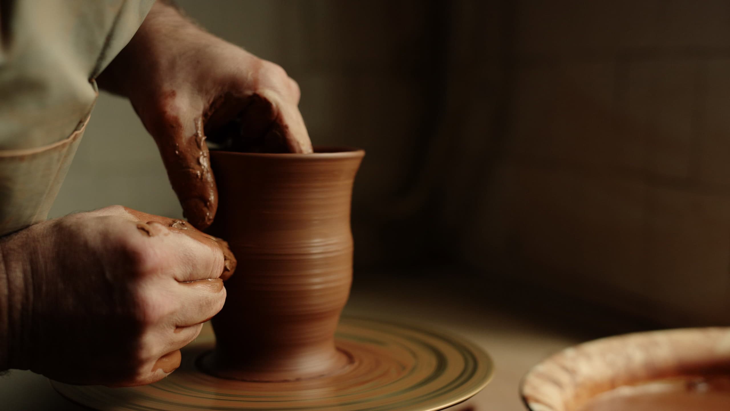 Unknown master forming jar from wet clay piece in pottery. Unrecognized clay artist modeling product on potters wheel in workshop. Closeup man hands sculpting in studio.