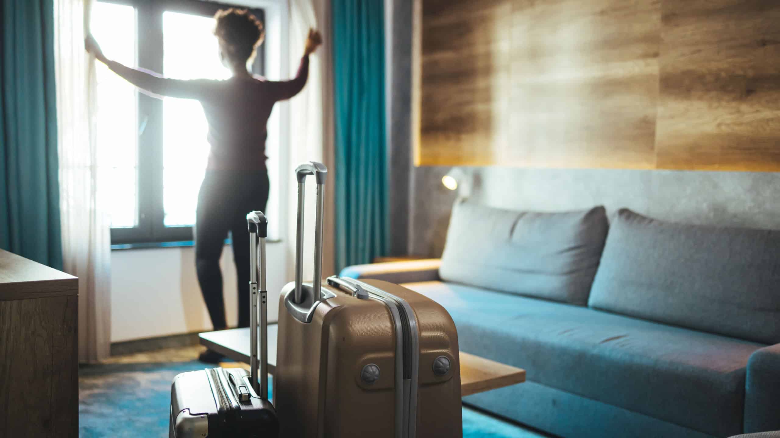 Luggage in modern hotel room with happy young adult female relaxing nearly window, African woman tourist looking to beautiful nature view. Time to travel, relaxation, journey, trip and vacation