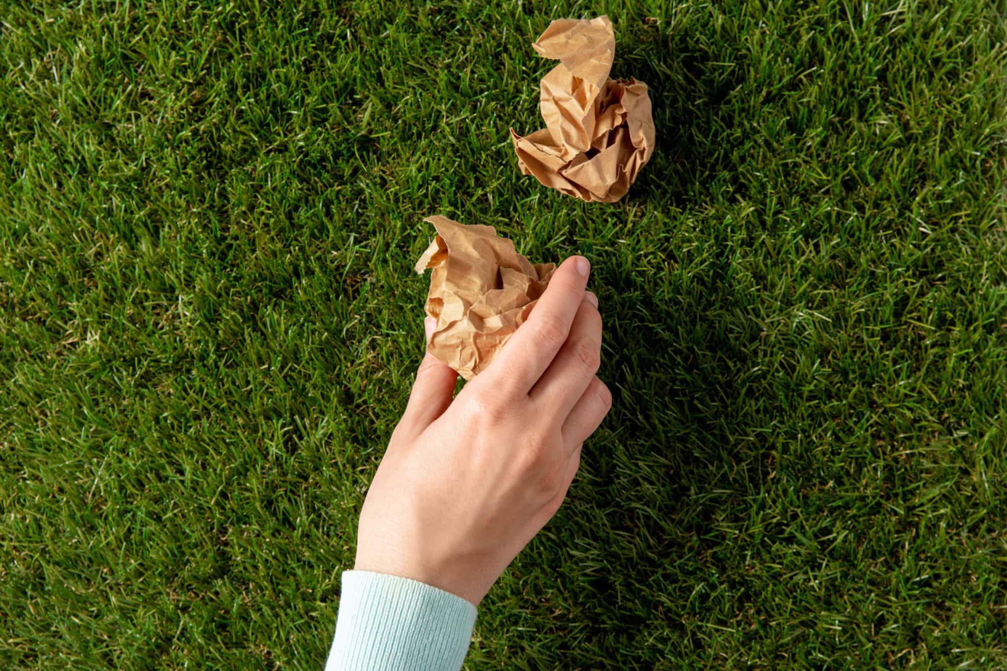 environment, volunteering and ecology concept - close up of hand picking crumpled paper waste from grass