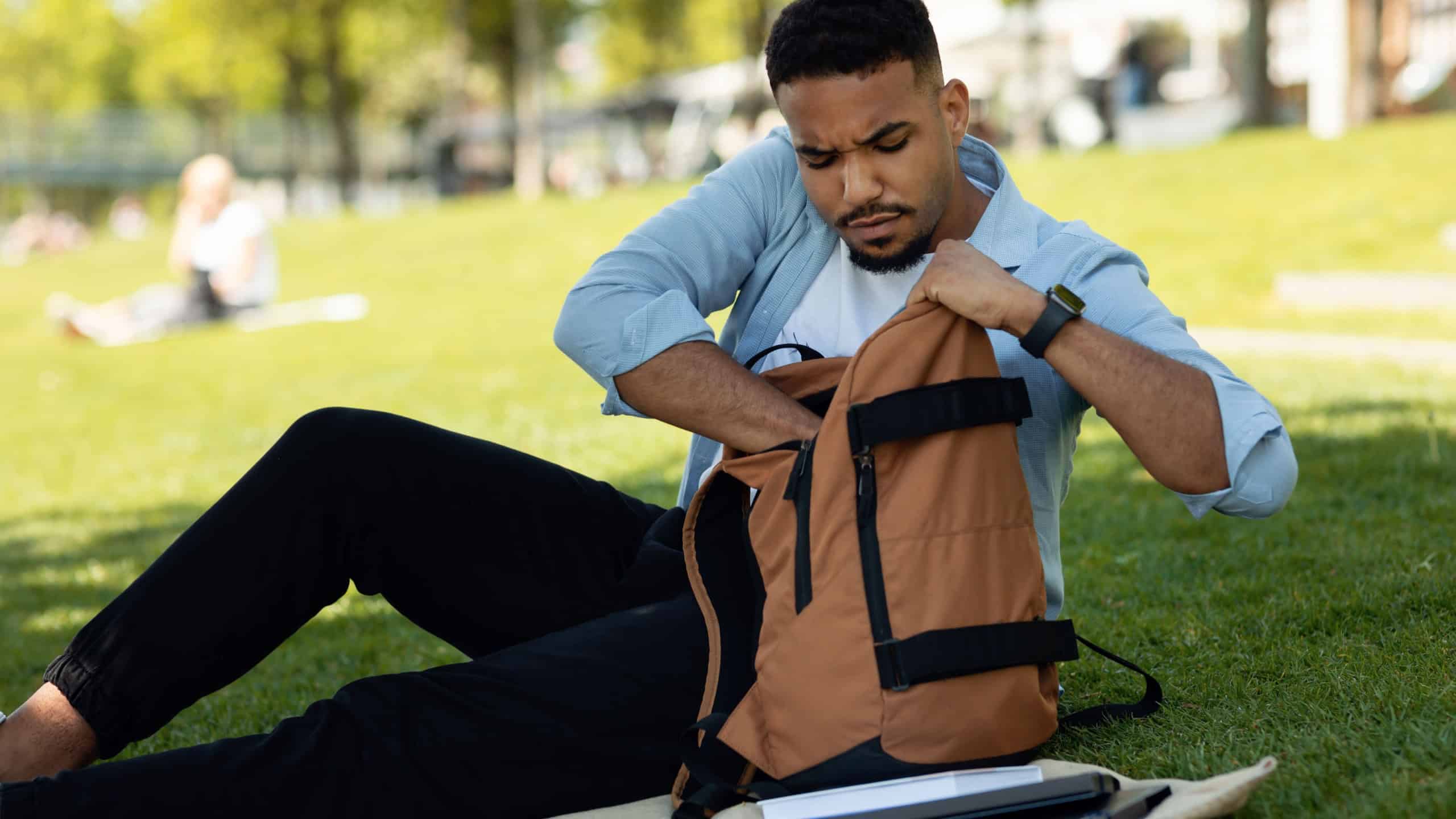 Serious african american guy looking inside his backpack and searching something, resting in city park outdoors. Black male student getting something out of his bag
