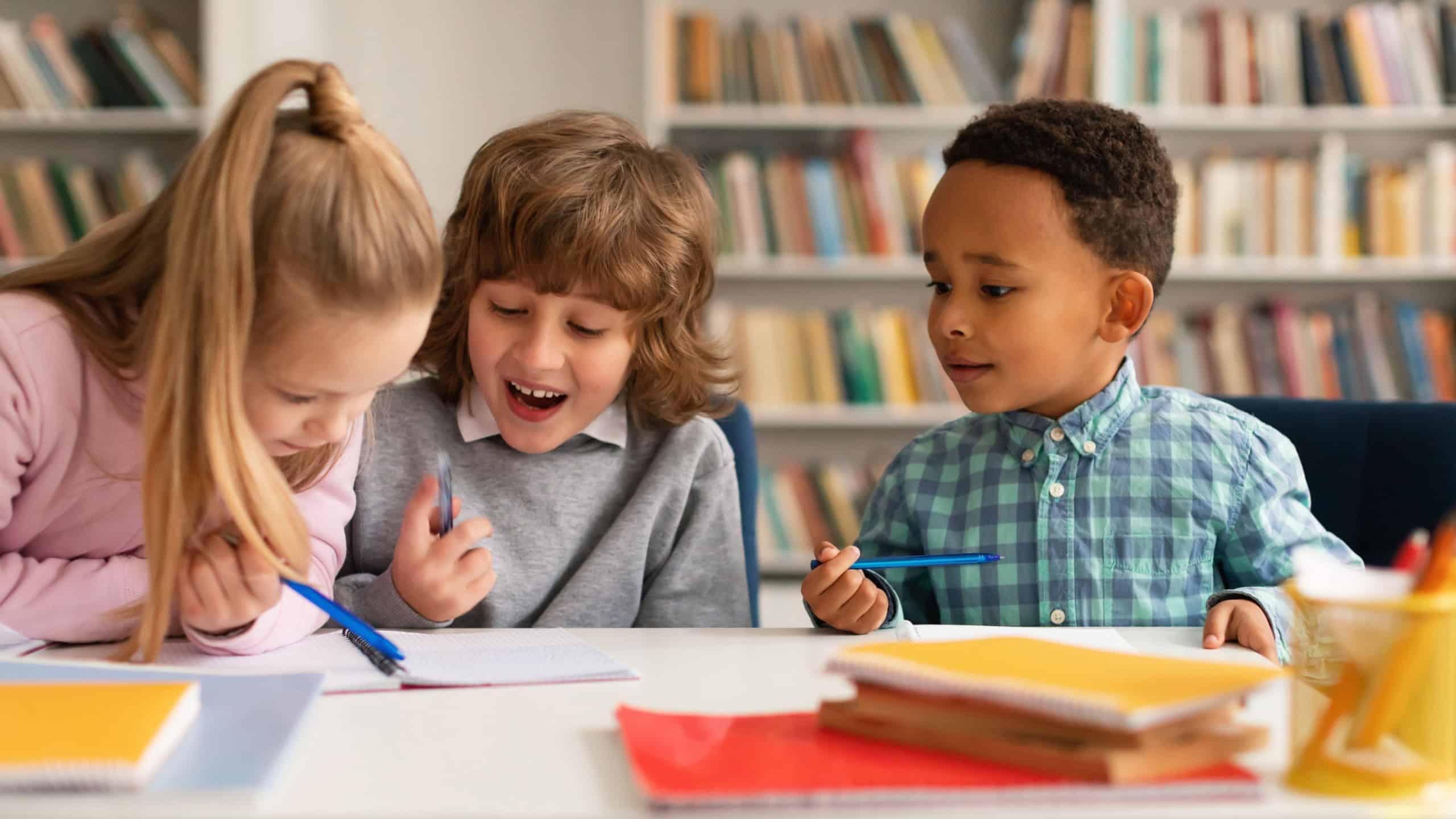 School time. Multiethnic kids writing in notebooks and smiling, talking with each other and laughing, sitting at table in classrom, studying in primary school
