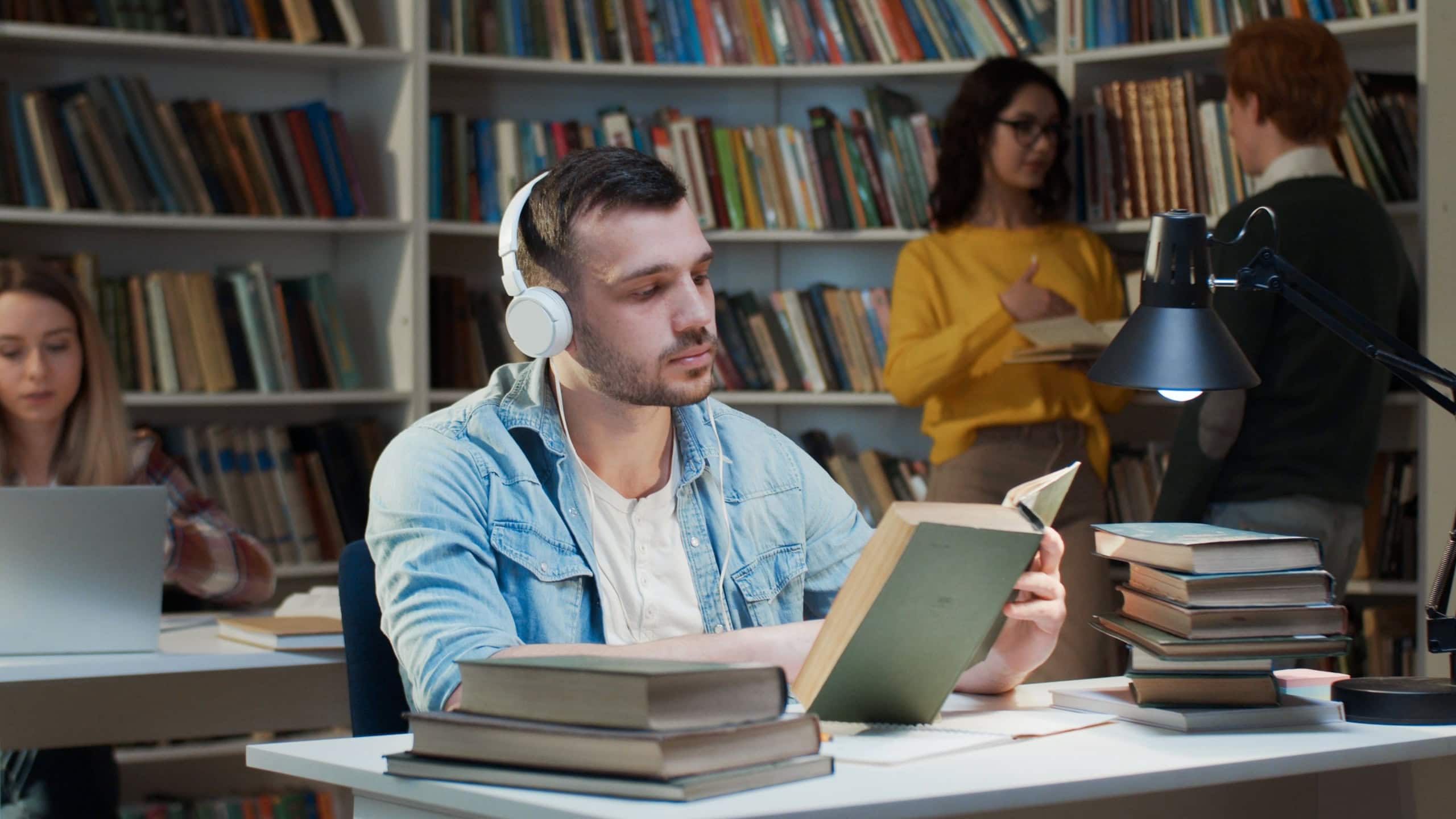 Caucasian young male student in headphones turning pages in old textbooks and studying. Listening to music in library. Guy reading book in bibliotheca. Education concept.